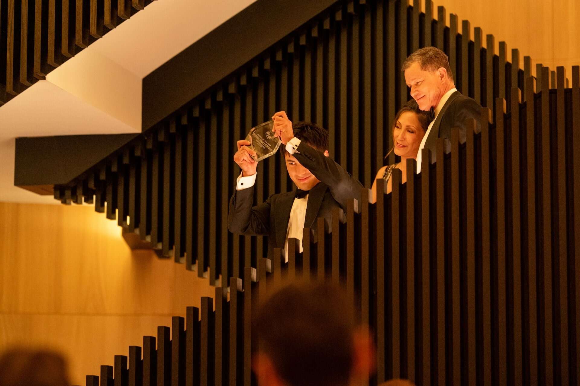 A man in a tuxedo holds up a glass award on a modern staircase, smiling, with a woman and another man in formal attire standing beside him, all appearing to celebrate.