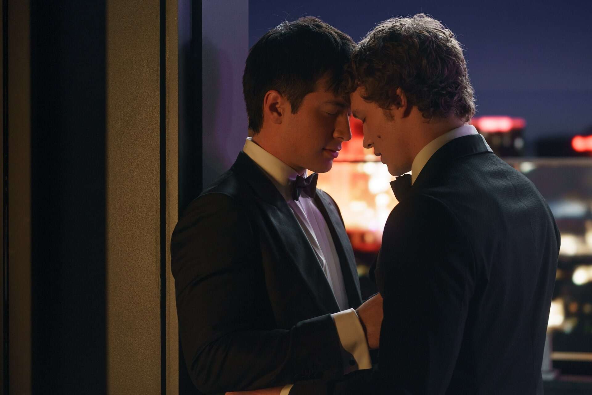 Two men in tuxedos stand close together, gently touching foreheads and holding hands, sharing an intimate moment by a window with city lights in the background.
