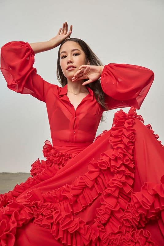 A woman wearing a vibrant red dress with ruffled details poses gracefully against a plain white background, with her arms raised and a confident expression on her face.
