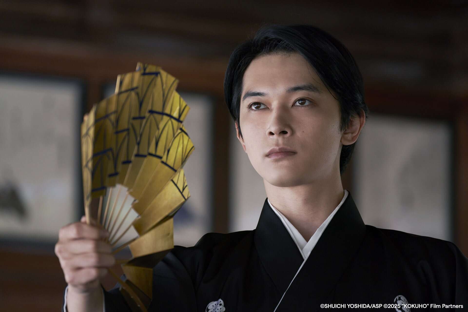 A young person in traditional Japanese attire holds a gold and white folding fan, looking forward with a serious expression. The background is softly blurred with framed artwork visible on the walls.