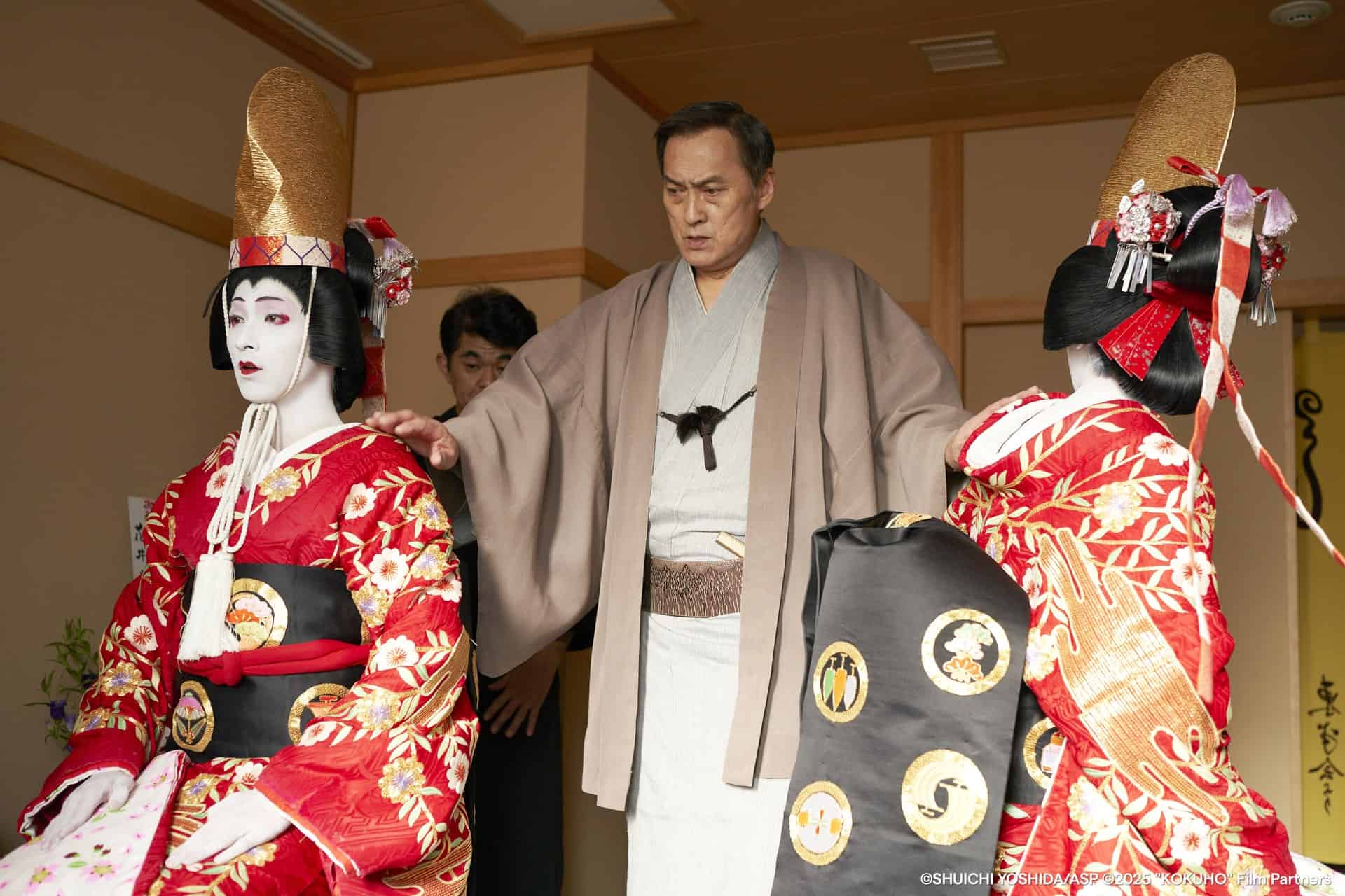 A man in traditional Japanese clothing stands between two people dressed as oiran courtesans in elaborate red kimonos and tall gold headdresses, indoors with wooden walls.