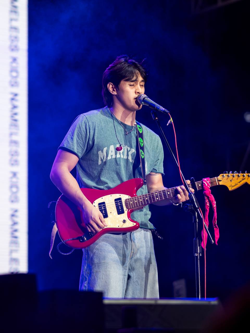 A musician in a blue MARIKINA t-shirt plays an electric guitar and sings into a microphone on stage, with blue lighting and a vertical NAMELESS KIDS sign in the background.
