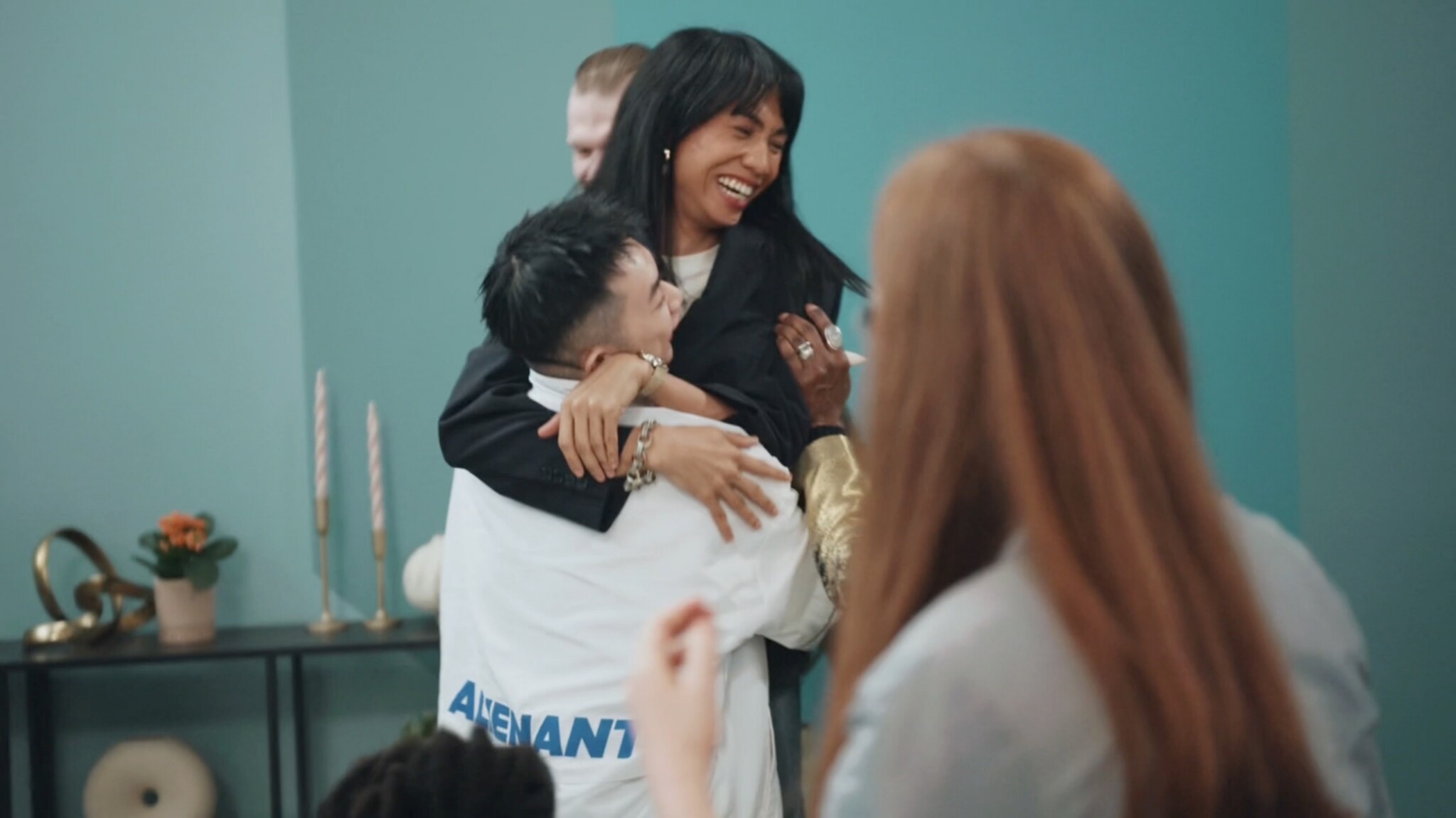 A woman laughs joyfully as she is lifted in a hug by a person in a white jacket, surrounded by smiling friends in a brightly lit room with teal walls.