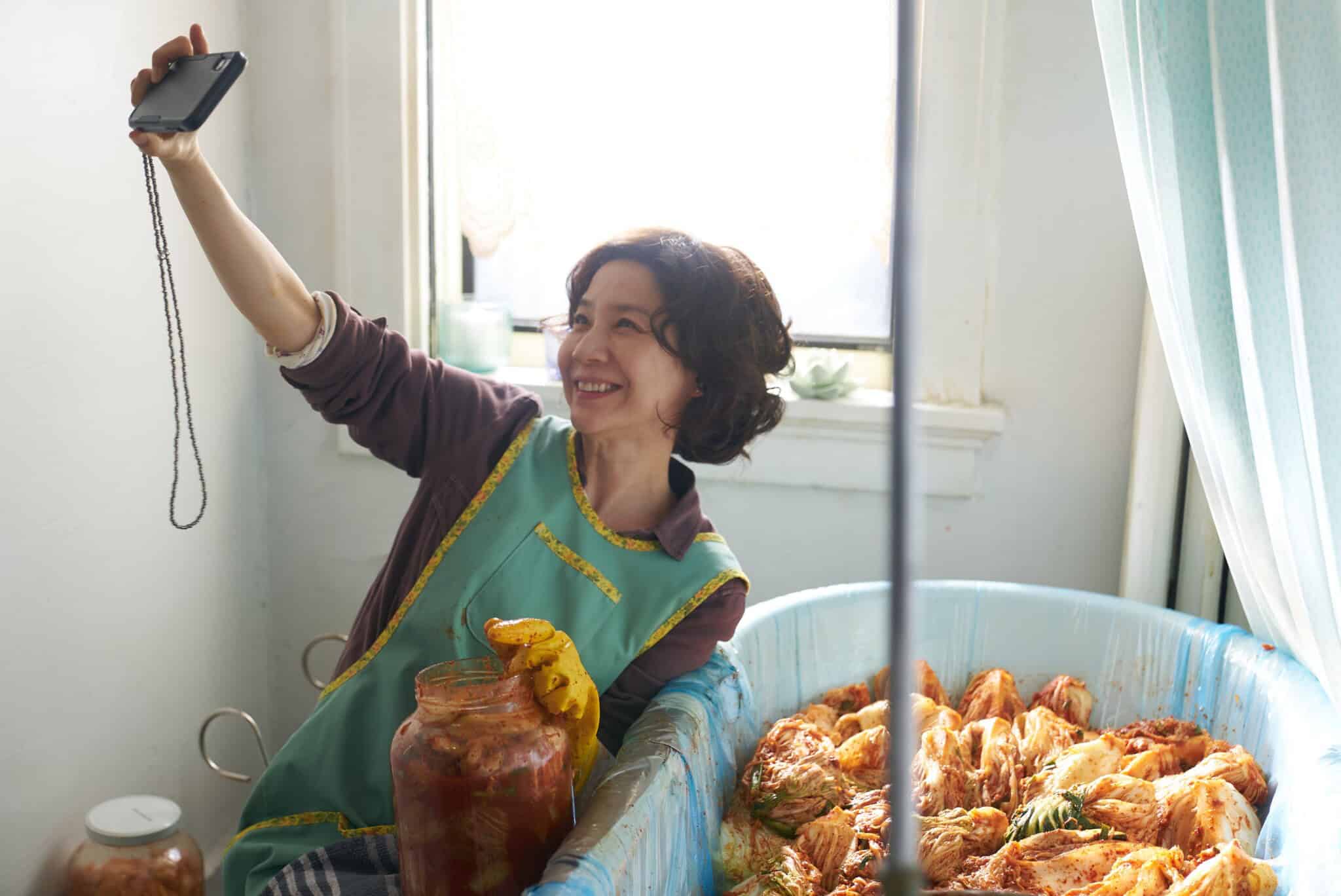 A woman wearing an apron and yellow glove smiles and takes a selfie while sitting next to a large tub of kimchi in a bright room, holding a jar filled with kimchi.