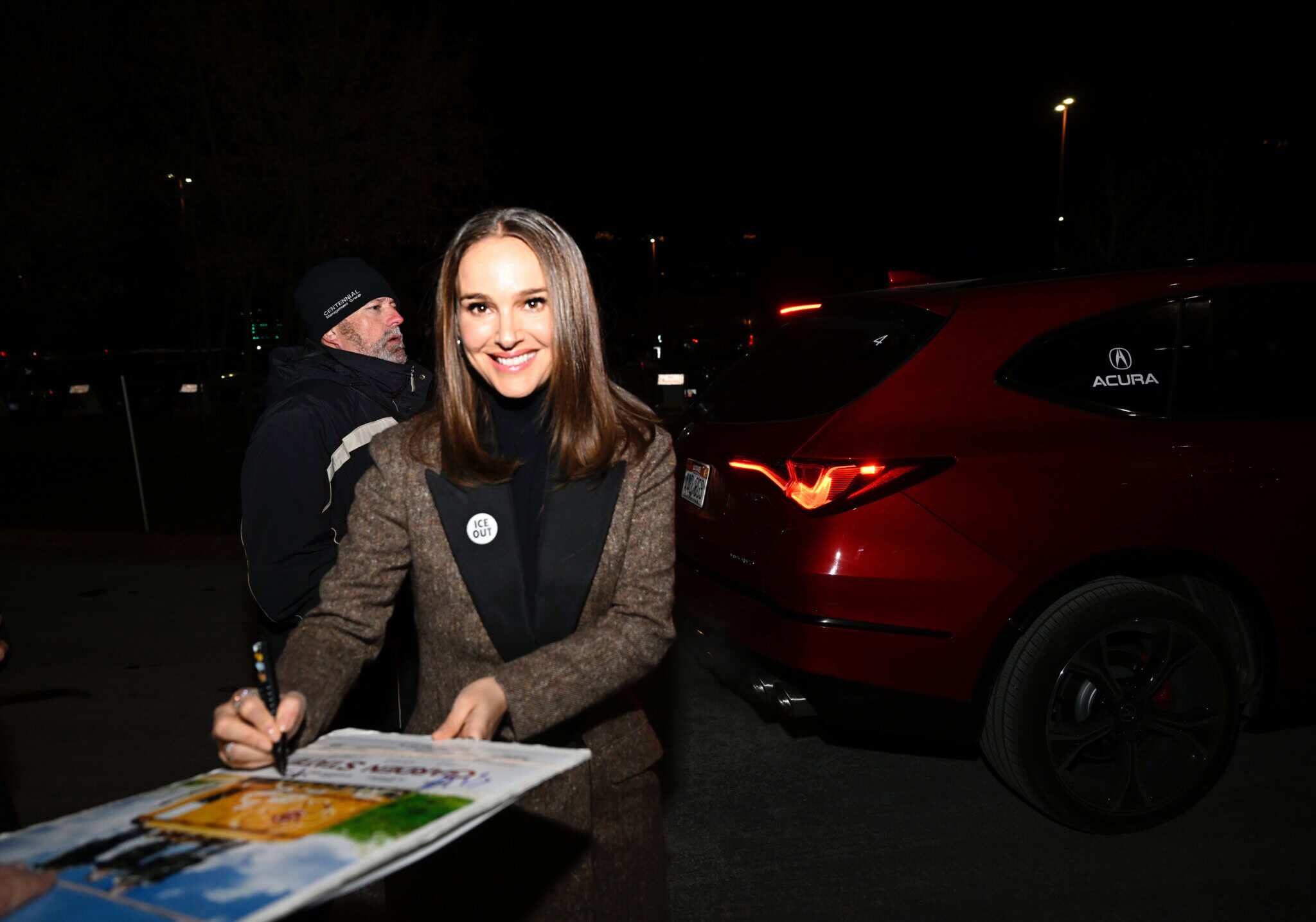 A woman with straight brown hair, wearing a brown coat over a black top, smiles as she signs a poster outdoors at night near a red Acura SUV. Another person stands in the background.