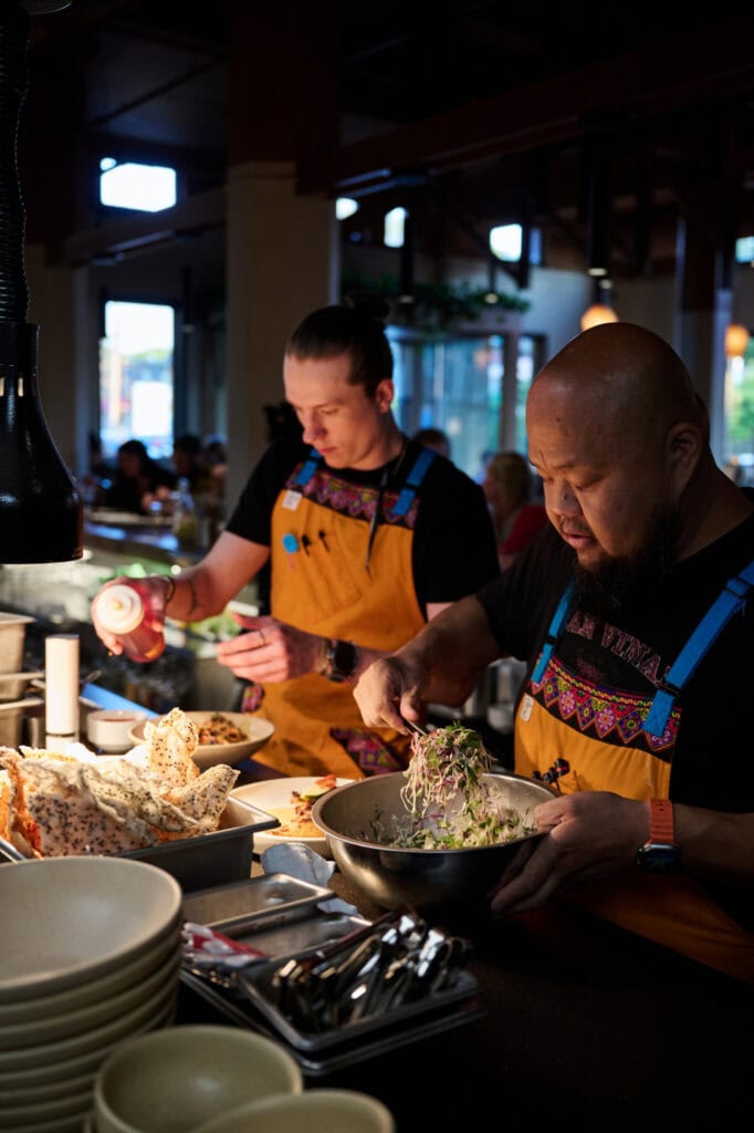 Two chefs wearing colorful aprons prepare food at a restaurant counter. One is plating a salad from a large bowl, while the other decorates a dish with sauce. Stacks of plates and food ingredients are visible nearby.