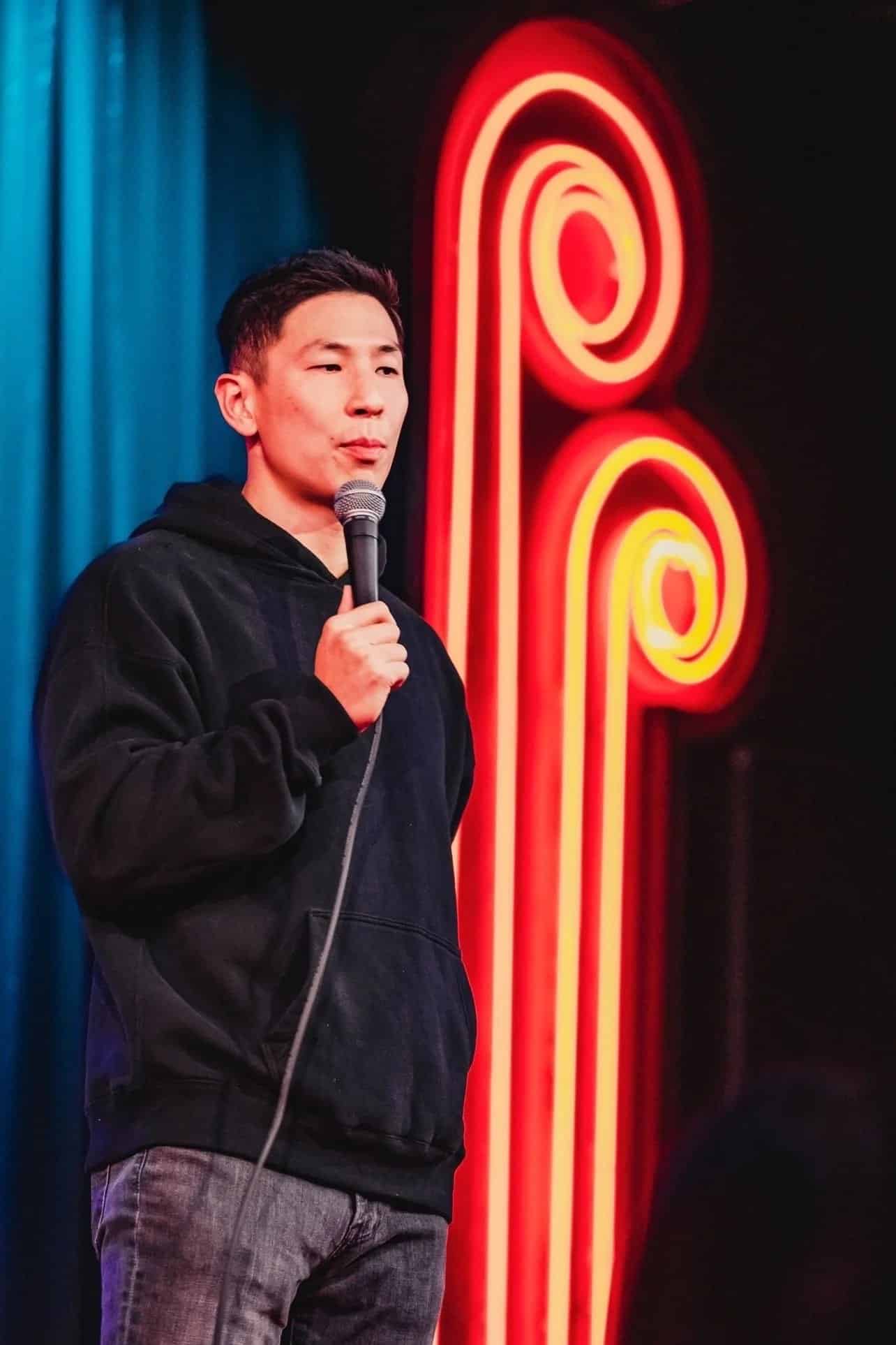 A person in a black hoodie stands on stage holding a microphone, performing stand-up comedy. Behind them is a red neon light design against a dark background with blue curtains.