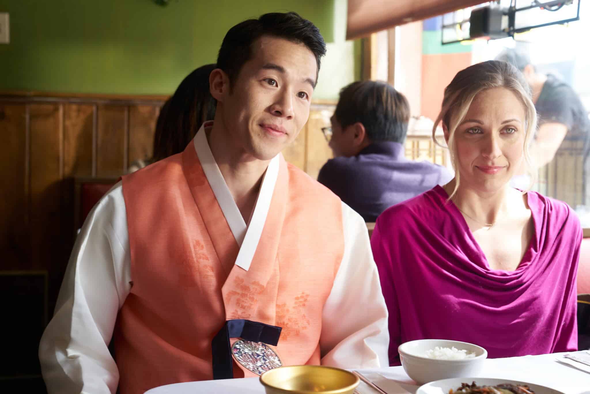 A man in a traditional Korean hanbok and a woman in a magenta top sit together at a restaurant table set with bowls and plates, with other diners visible in the background.