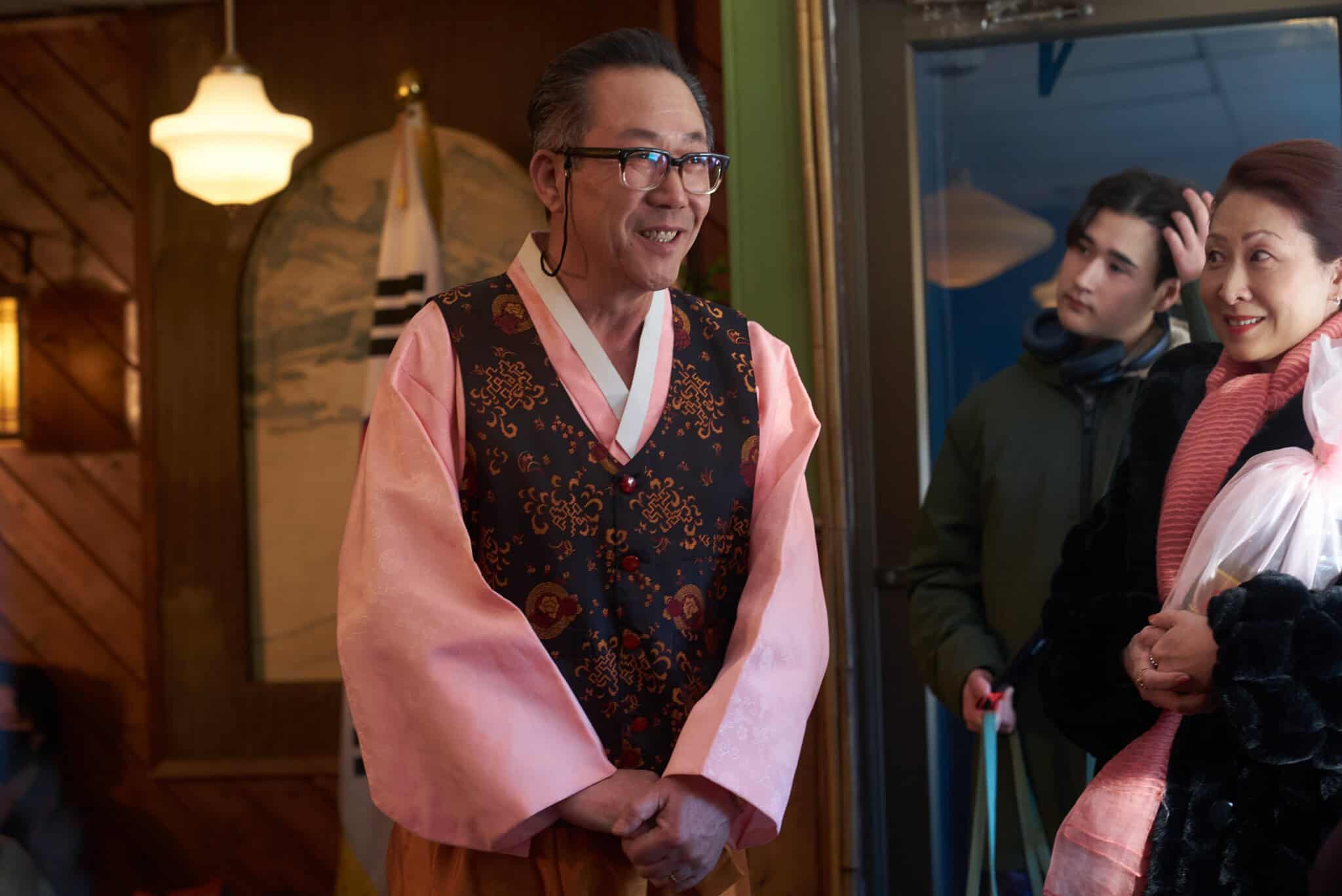 A man in traditional Korean clothing smiles and speaks indoors, while a woman in a pink scarf and a young man in a green jacket stand nearby, listening. Warm lighting creates a cozy atmosphere.