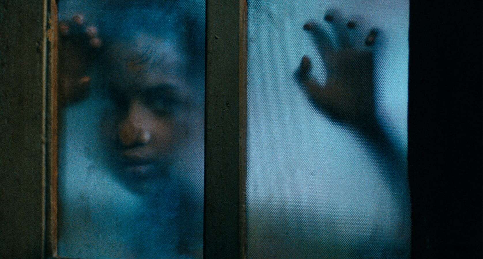 A person presses their face and hands against a foggy glass window, with features blurred and a somber expression visible through the translucent surface, evoking the cinematic poignancy of a Mira Nair film.