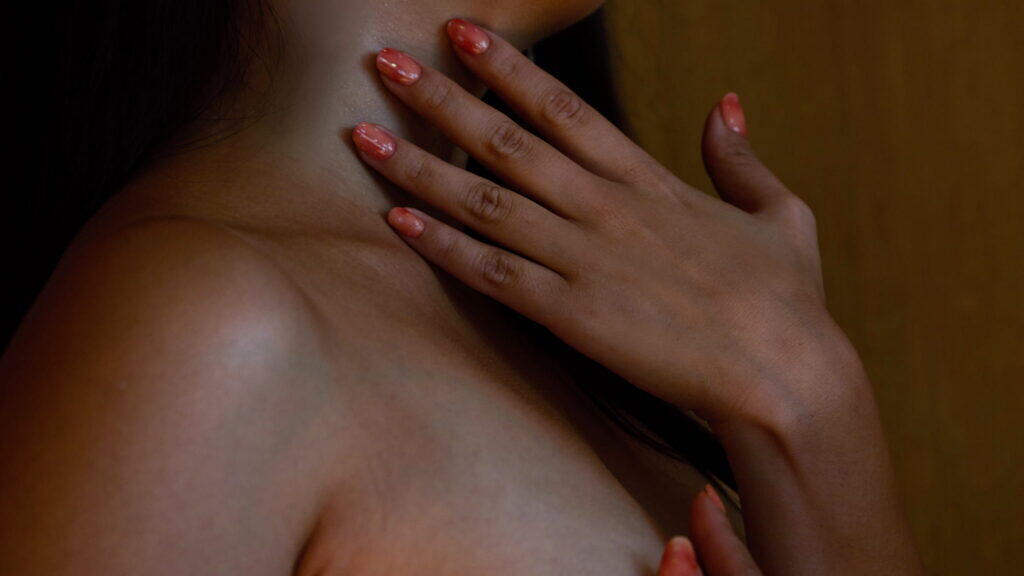 A close-up of a persons hand with glossy, peach-colored nails resting gently on their bare chest and neck, highlighting smooth skin and painted fingernails.