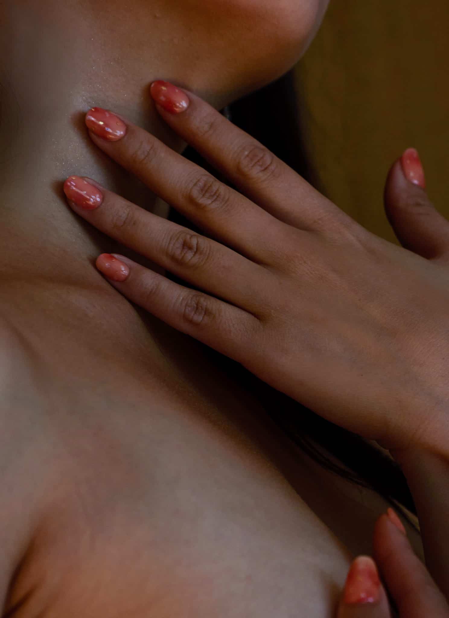 A close-up of a persons hand with glossy, peach-colored nails resting gently on their bare chest and neck, highlighting smooth skin and painted fingernails.