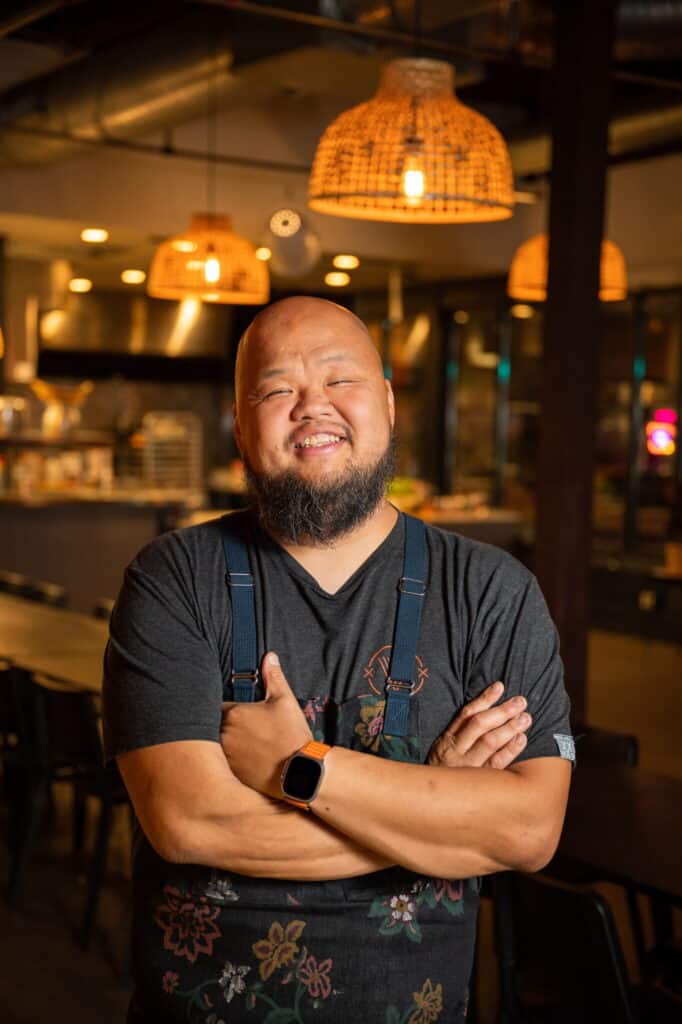 A smiling man with a beard stands in a warmly lit restaurant, wearing a dark shirt, blue suspenders, and a smartwatch, with his arms folded across his chest. Woven pendant lights and kitchen equipment are visible in the background.