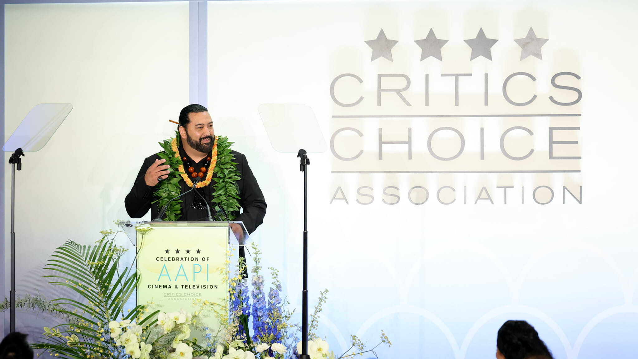A man wearing a large green lei speaks at a podium decorated with flowers at a Critics Choice Association event celebrating AAPI cinema and television. The background features the event’s logo and name in large letters.