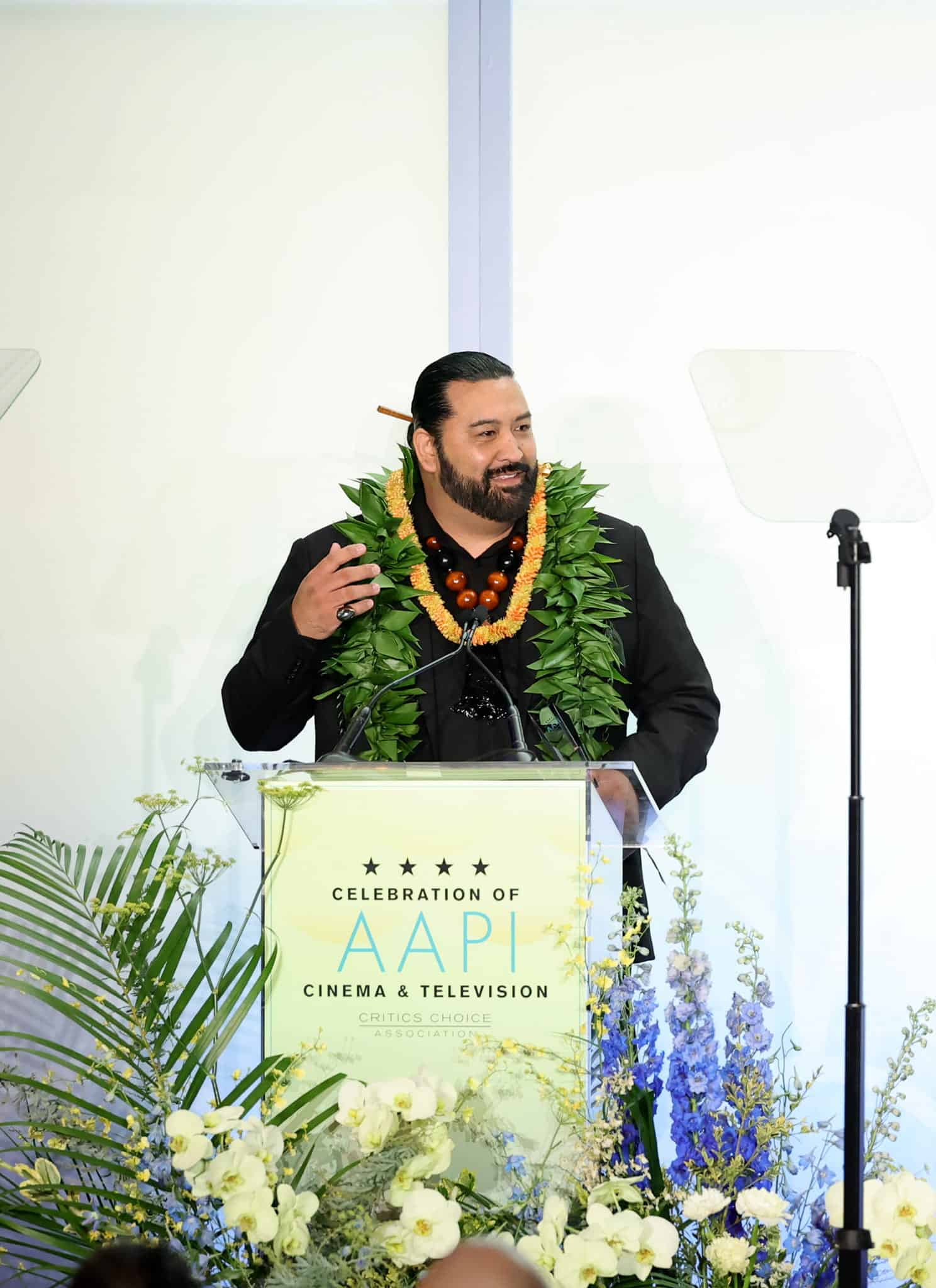 A man wearing a large green lei speaks at a podium decorated with flowers at a Critics Choice Association event celebrating AAPI cinema and television. The background features the event’s logo and name in large letters.