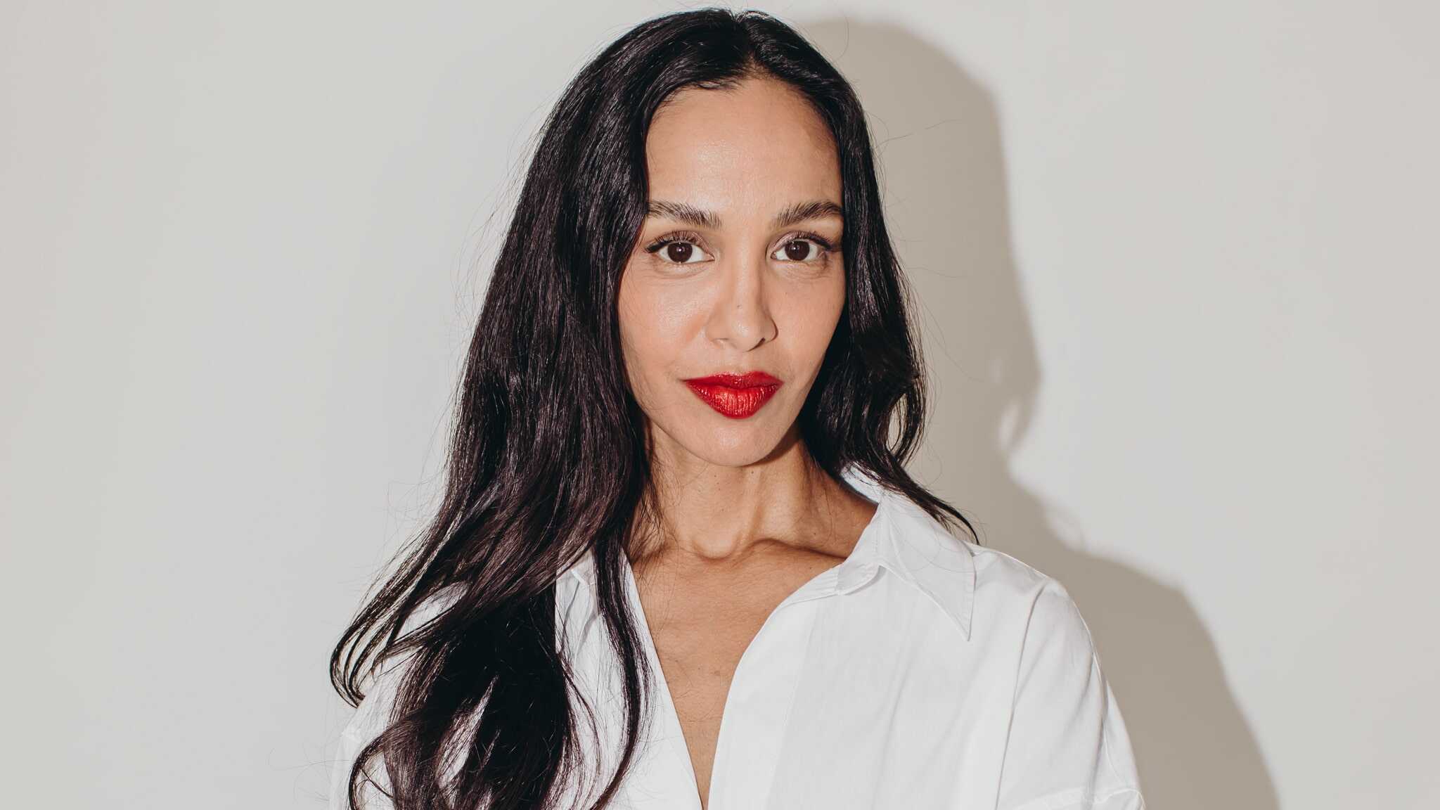 A woman with long dark hair and red lipstick wearing a white button-up shirt stands against a plain white background, looking directly at the camera with a neutral expression.