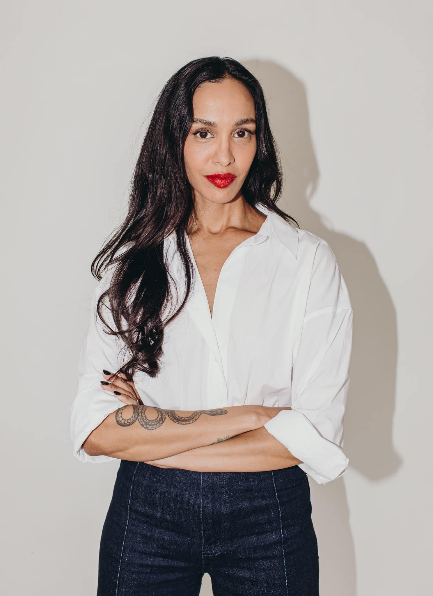 A woman with long dark hair and red lipstick wearing a white button-up shirt stands against a plain white background, looking directly at the camera with a neutral expression.