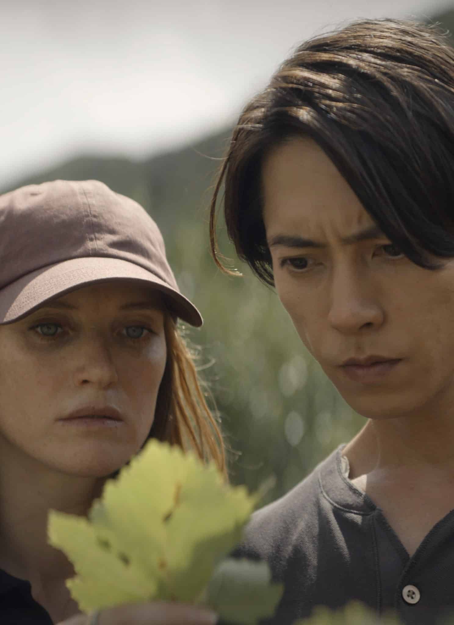 A woman in a baseball cap and a man stand closely together outdoors, intently examining green leaves on a plant, with greenery and blurred natural scenery in the background.