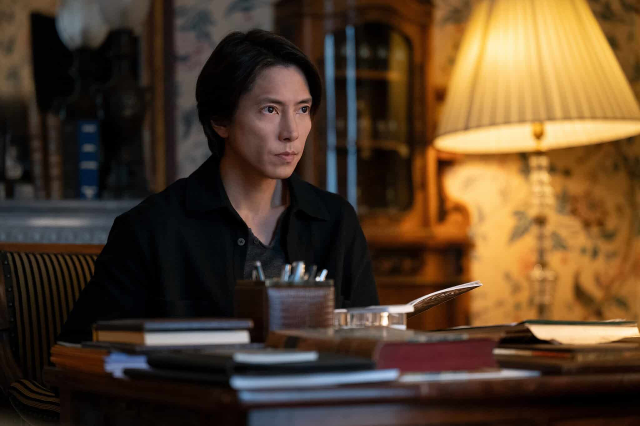 A person with dark hair sits at a desk cluttered with books and papers, looking serious. A lamp and wooden cabinet are in the softly lit, vintage-style background.