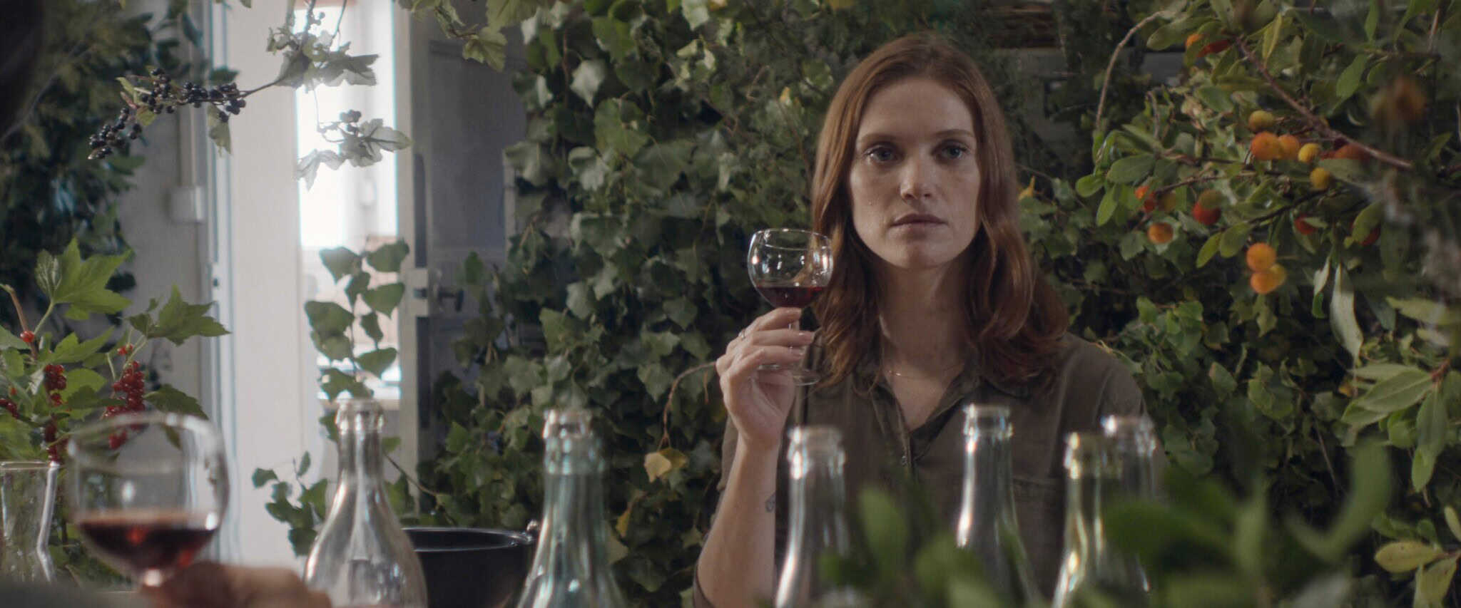 A woman with long brown hair holds a glass of red wine, sitting at a table with bottles, surrounded by lush green foliage and orange berries. She looks ahead with a neutral expression.