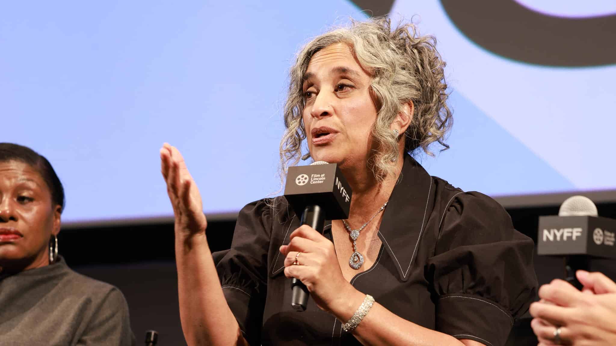 A woman with curly gray hair speaks into a microphone at an event, gesturing with her hand. She wears a dark dress and a pendant necklace. Another person and microphones labeled “NYFF” are visible.