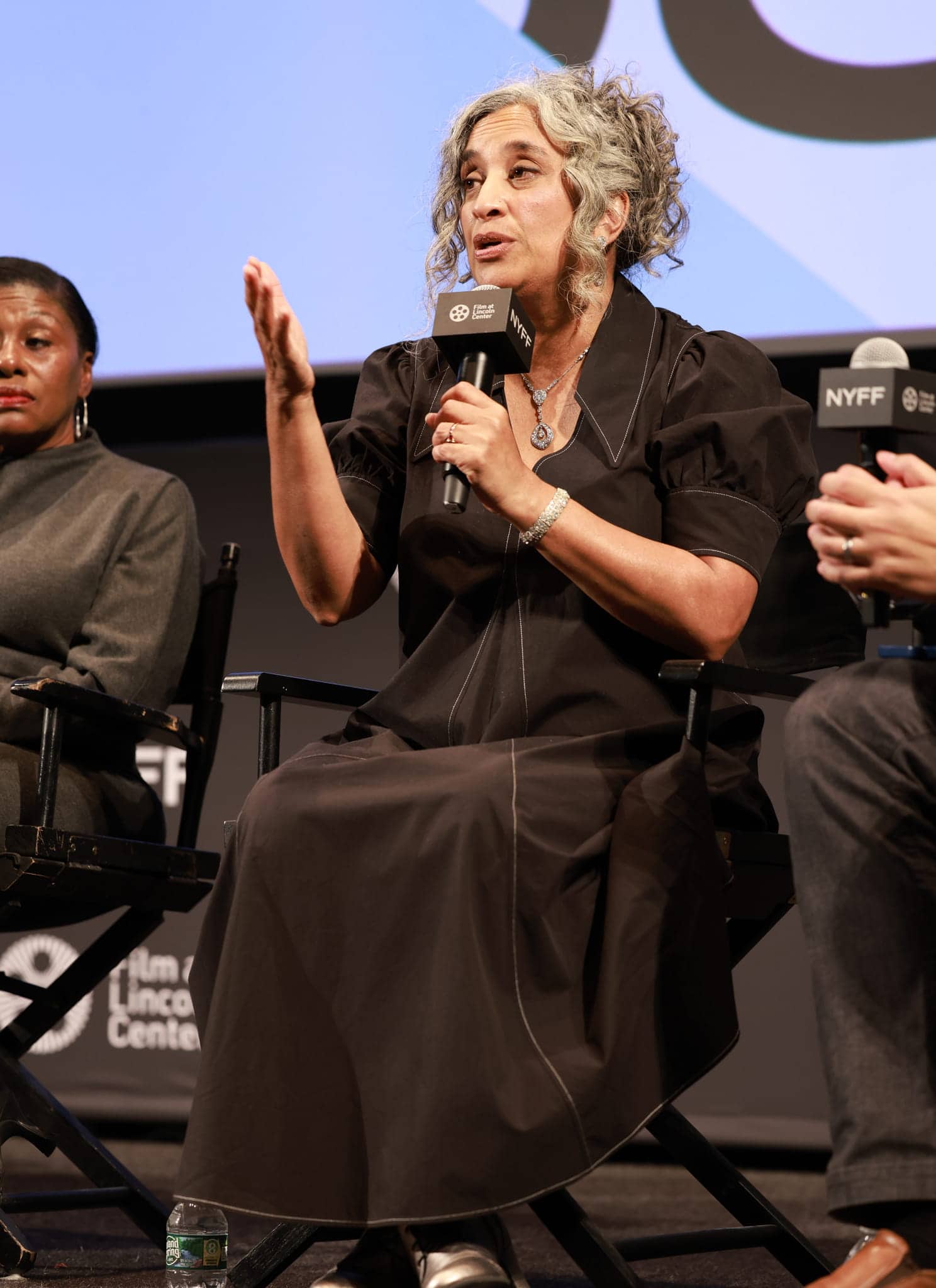 A woman with curly gray hair speaks into a microphone at an event, gesturing with her hand. She wears a dark dress and a pendant necklace. Another person and microphones labeled “NYFF” are visible.