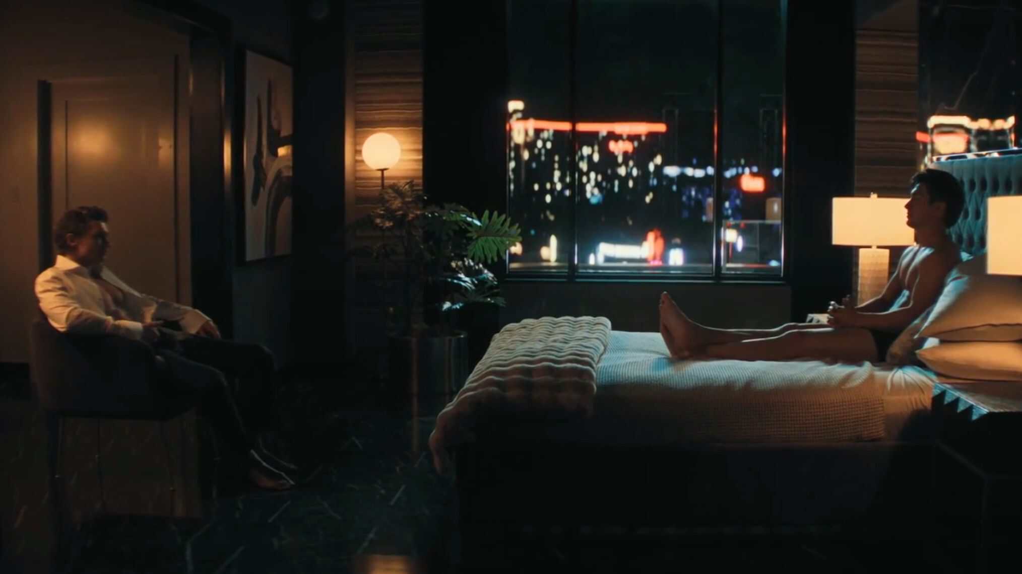 A dimly lit room with a bed in the center; one person sits fully clothed in a chair on the left while another person sits nude on the edge of the bed. City lights are visible through a large window in the background.