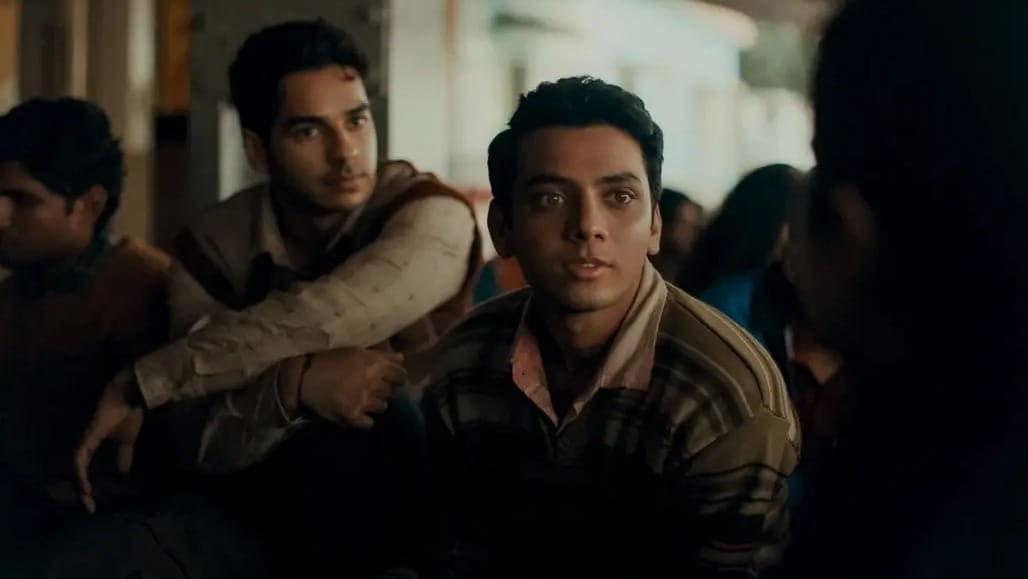 Two young men sit closely together in a group setting, looking attentively towards a person outside the frame. They are dressed in casual, warm clothing. The background is softly blurred, suggesting an indoor environment.