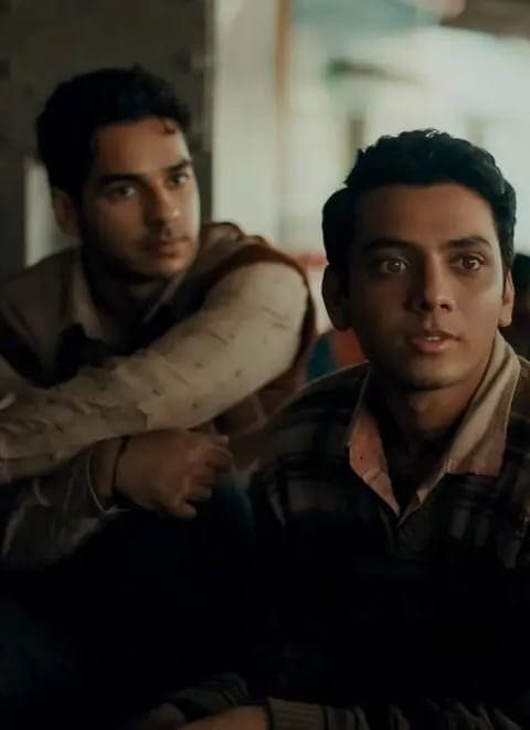 Two young men sit closely together in a group setting, looking attentively towards a person outside the frame. They are dressed in casual, warm clothing. The background is softly blurred, suggesting an indoor environment.