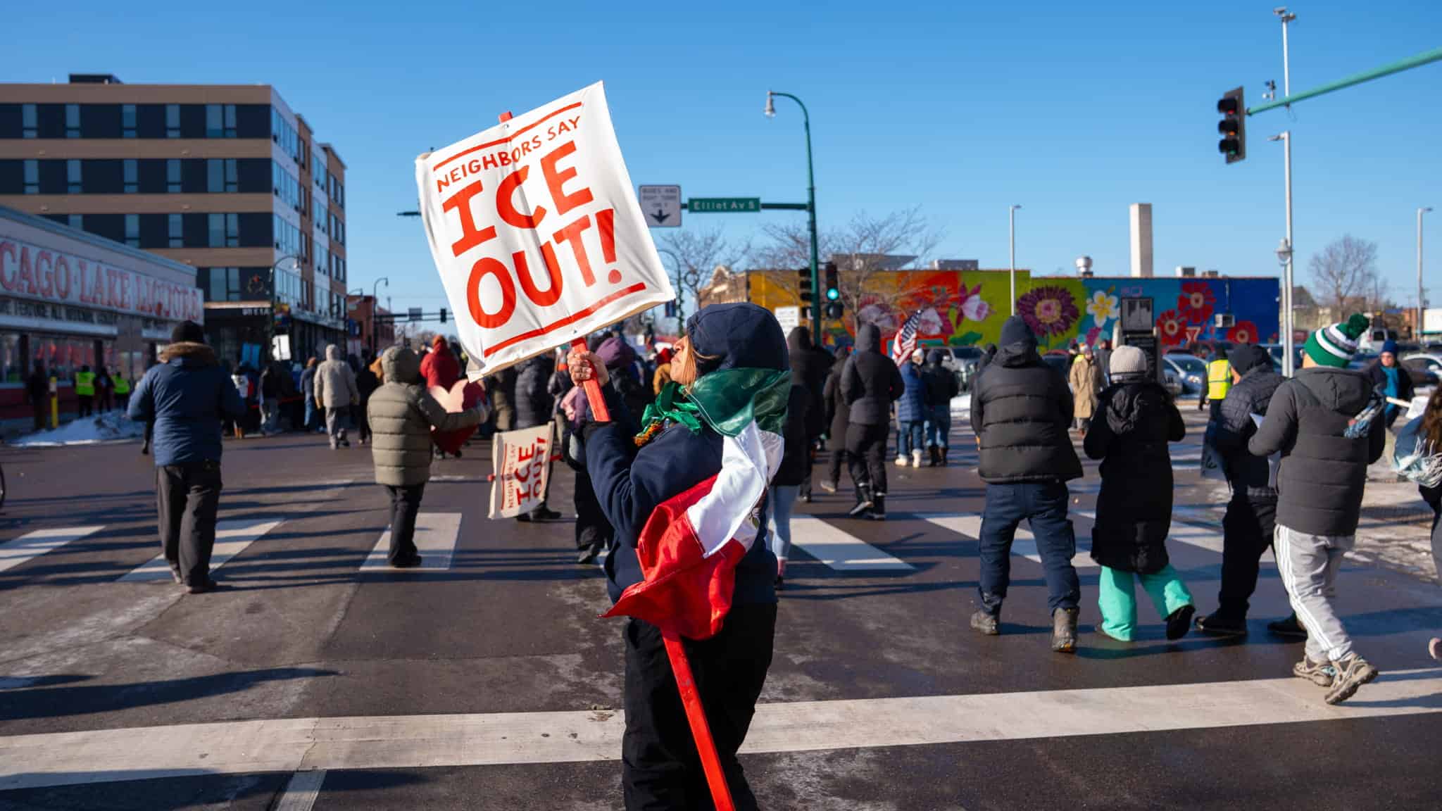 A person holding a sign that reads Neighbors Say ICE OUT! and draped in a flag stands in a city street among a group of people during a protest on a sunny winter day.