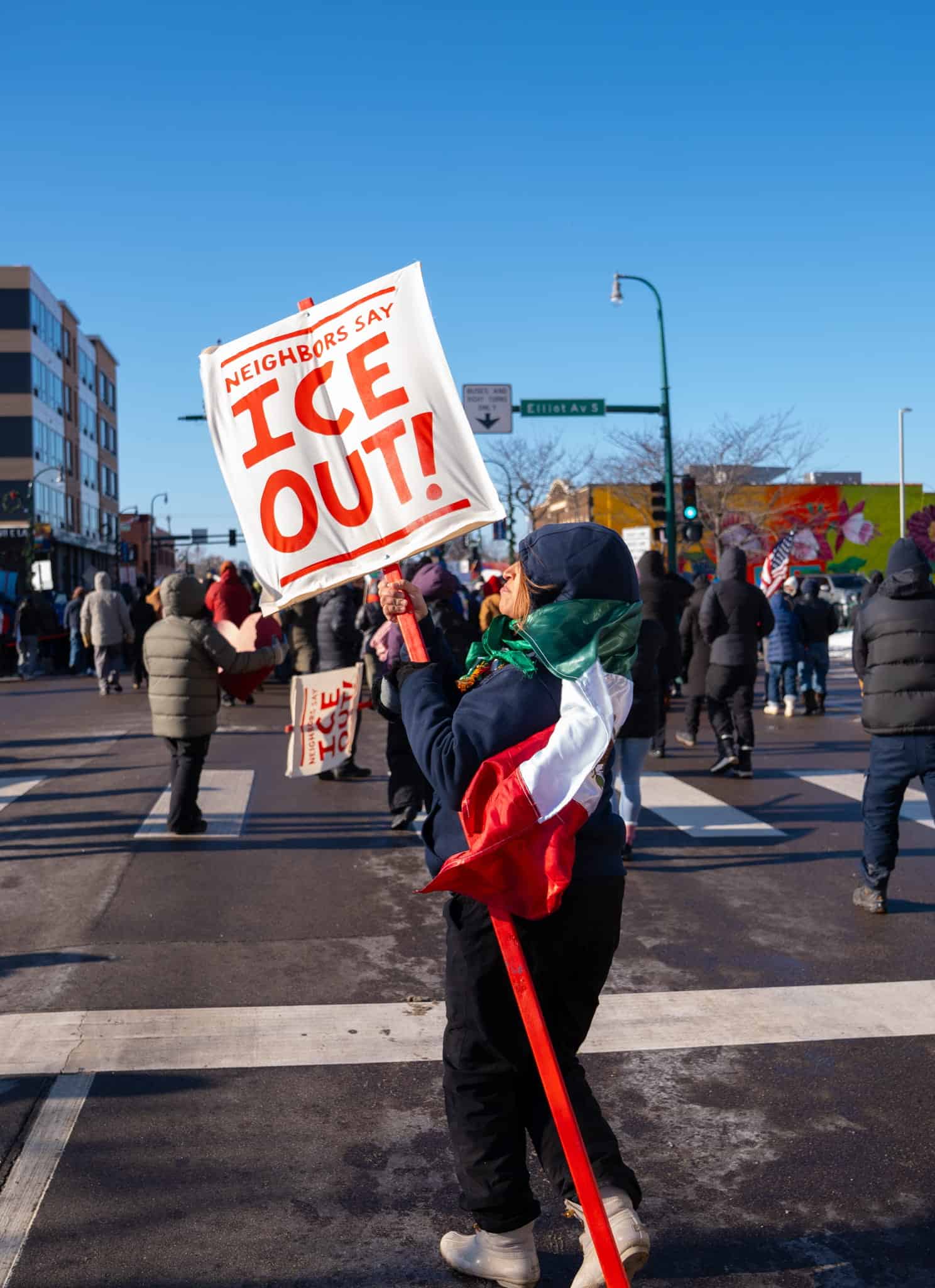 A person holding a sign that reads Neighbors Say ICE OUT! and draped in a flag stands in a city street among a group of people during a protest on a sunny winter day.