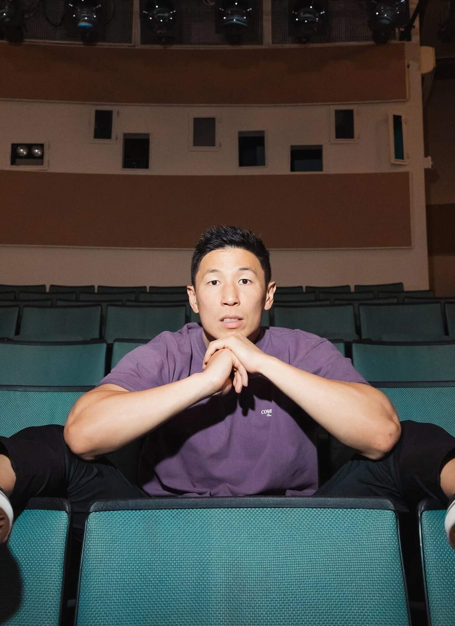 A person in a purple shirt sits in theater seats with legs stretched wide, feet on the armrests, and hands resting under their chin, looking directly at the camera in an empty auditorium.