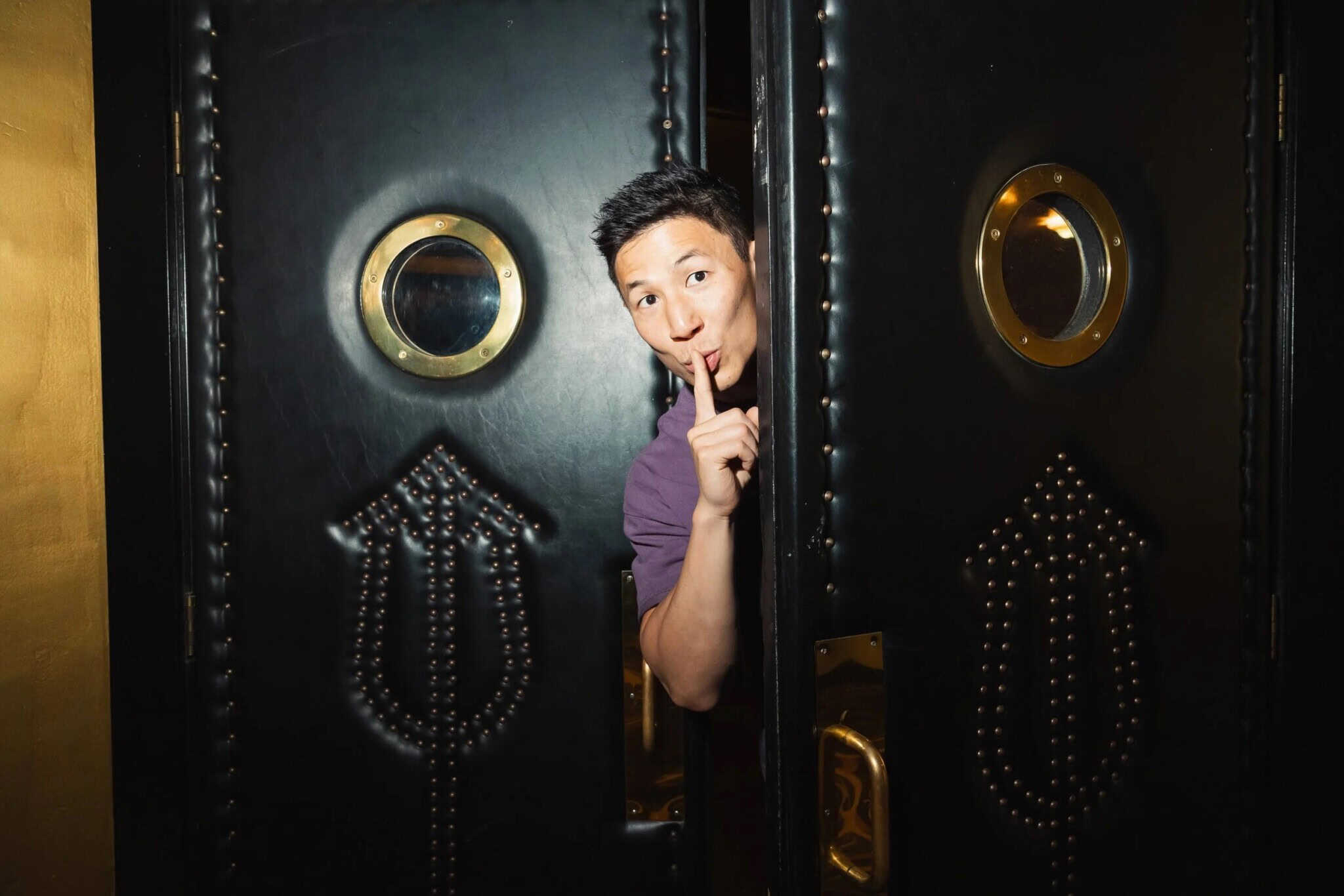 A man stands between two ornate black doors, holding a finger to his lips in a “shh” gesture, partially hidden while peeking out through the narrow opening.