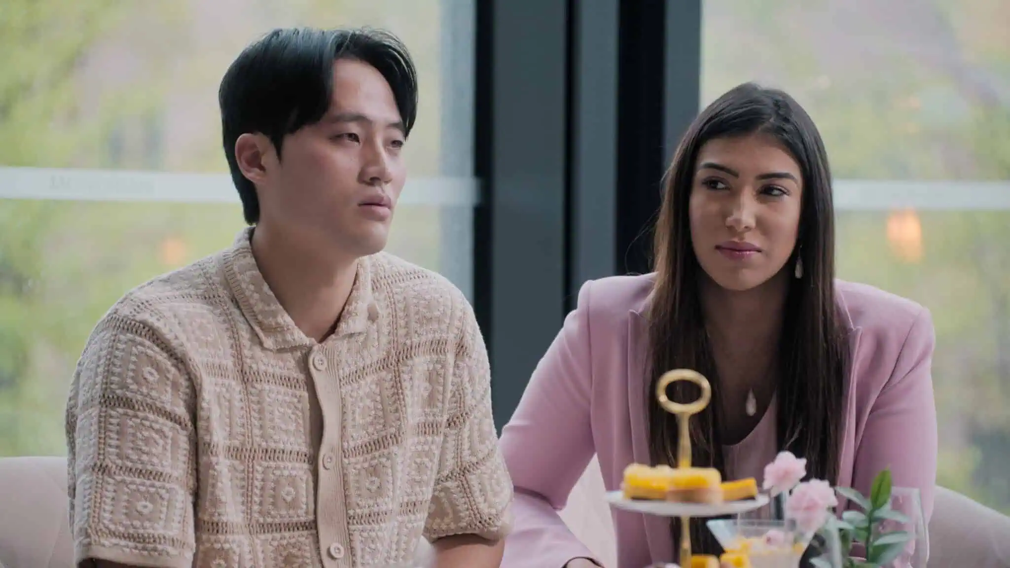 A man in a beige patterned shirt and a woman in a light pink blazer sit together indoors at a table with yellow desserts and flowers, looking thoughtfully to the side. Large windows show greenery outside.