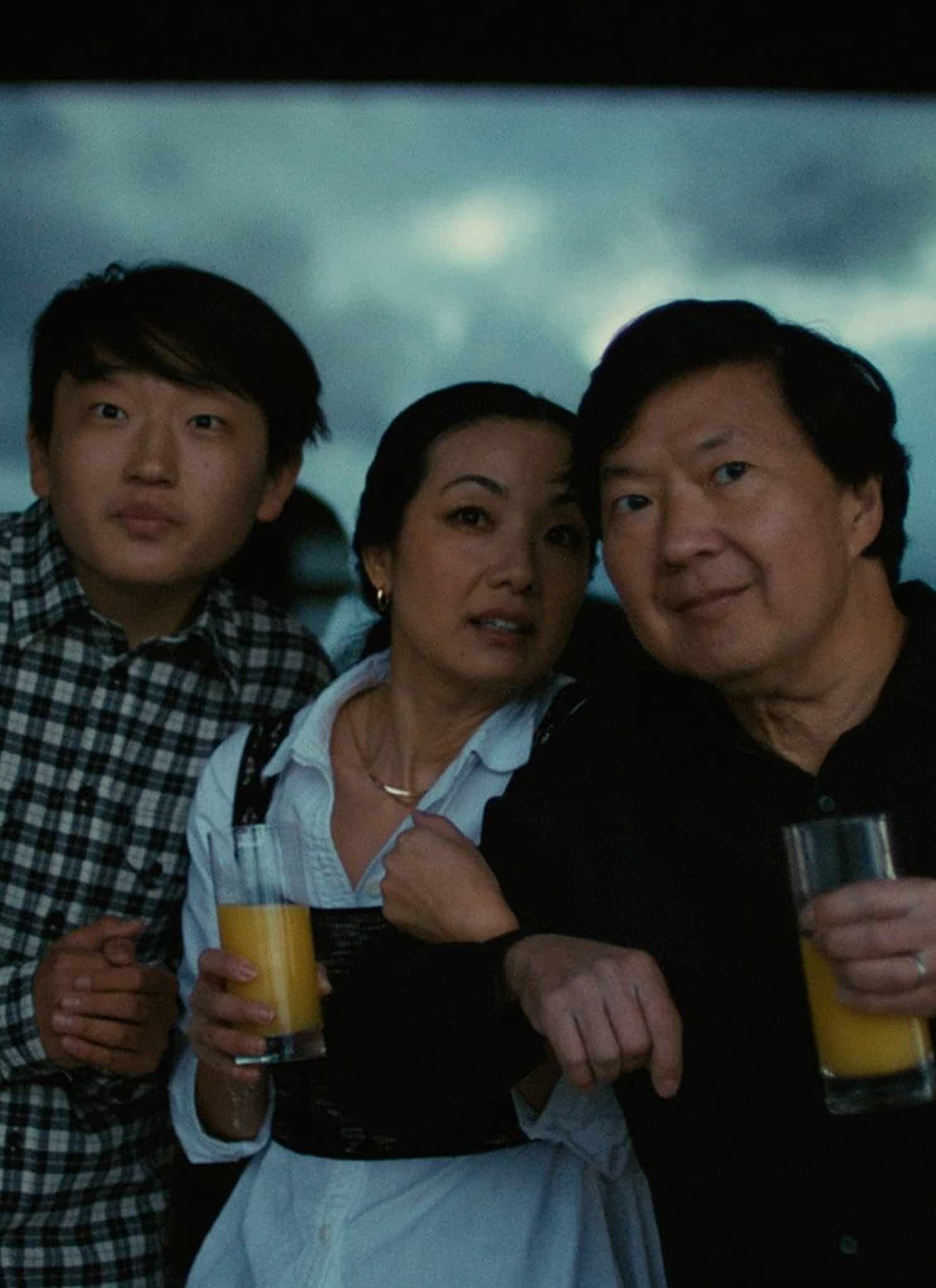 Three people stand close together in front of a window with a dark, cloudy sky in the background. Two of them hold glasses of orange juice and all appear to be looking at something off-camera.