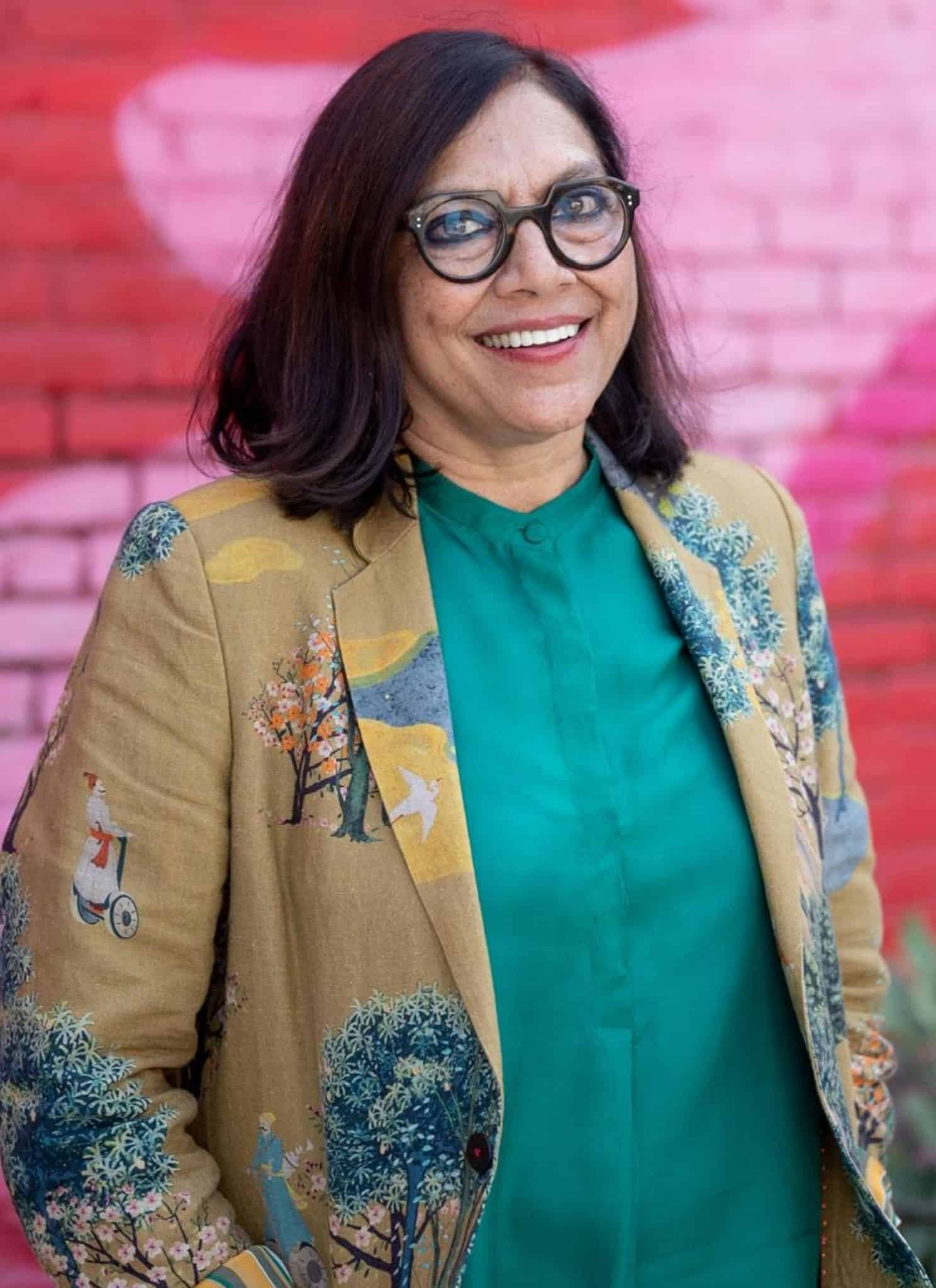 A woman with shoulder-length dark hair and glasses smiles while standing in front of a red brick wall with pink graffiti. She is wearing a green shirt and a tan jacket with colorful embroidery.