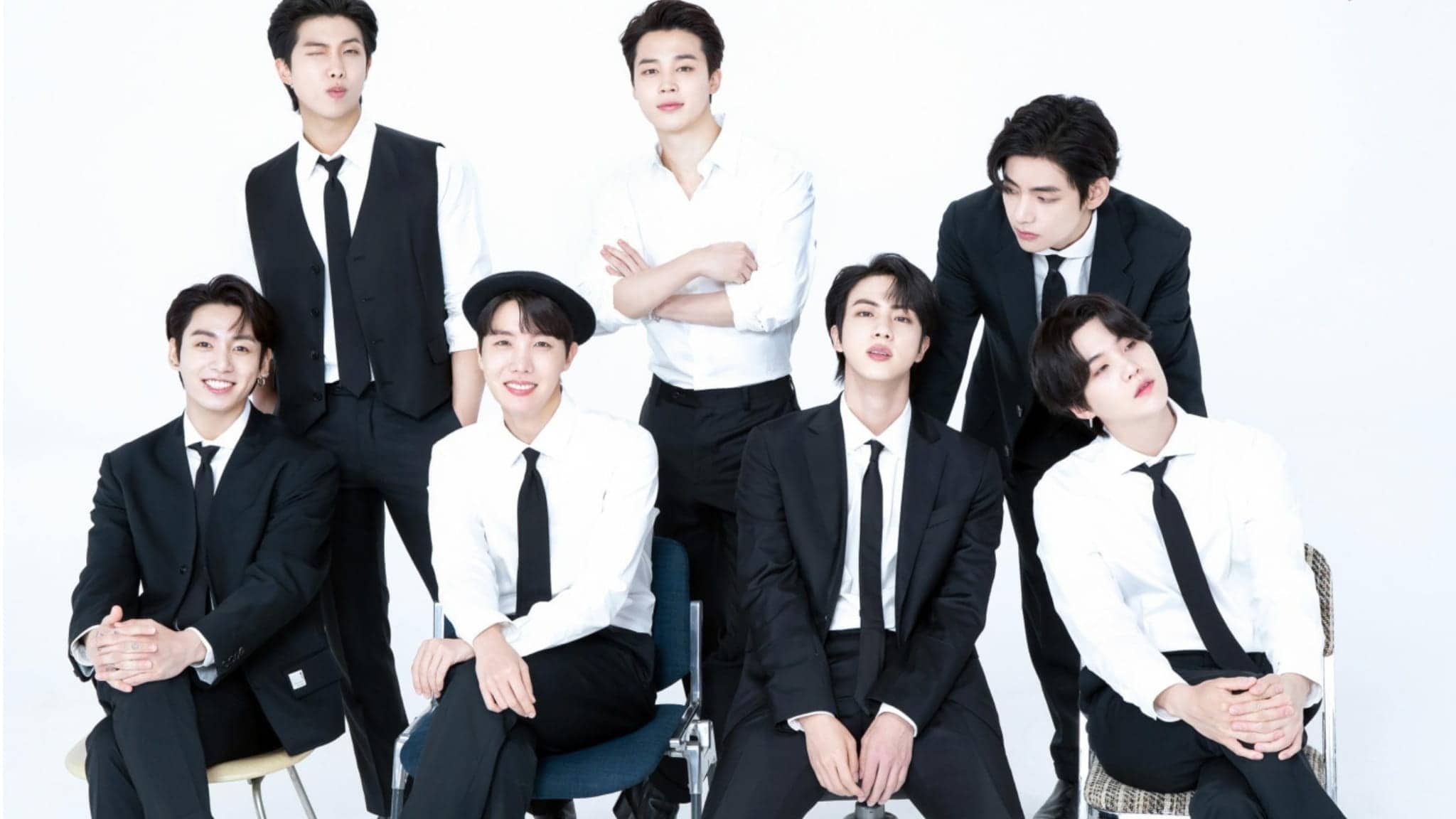 Seven young men dressed in black suits and white shirts pose together against a plain white background, some sitting and some standing, all smiling or looking relaxed.