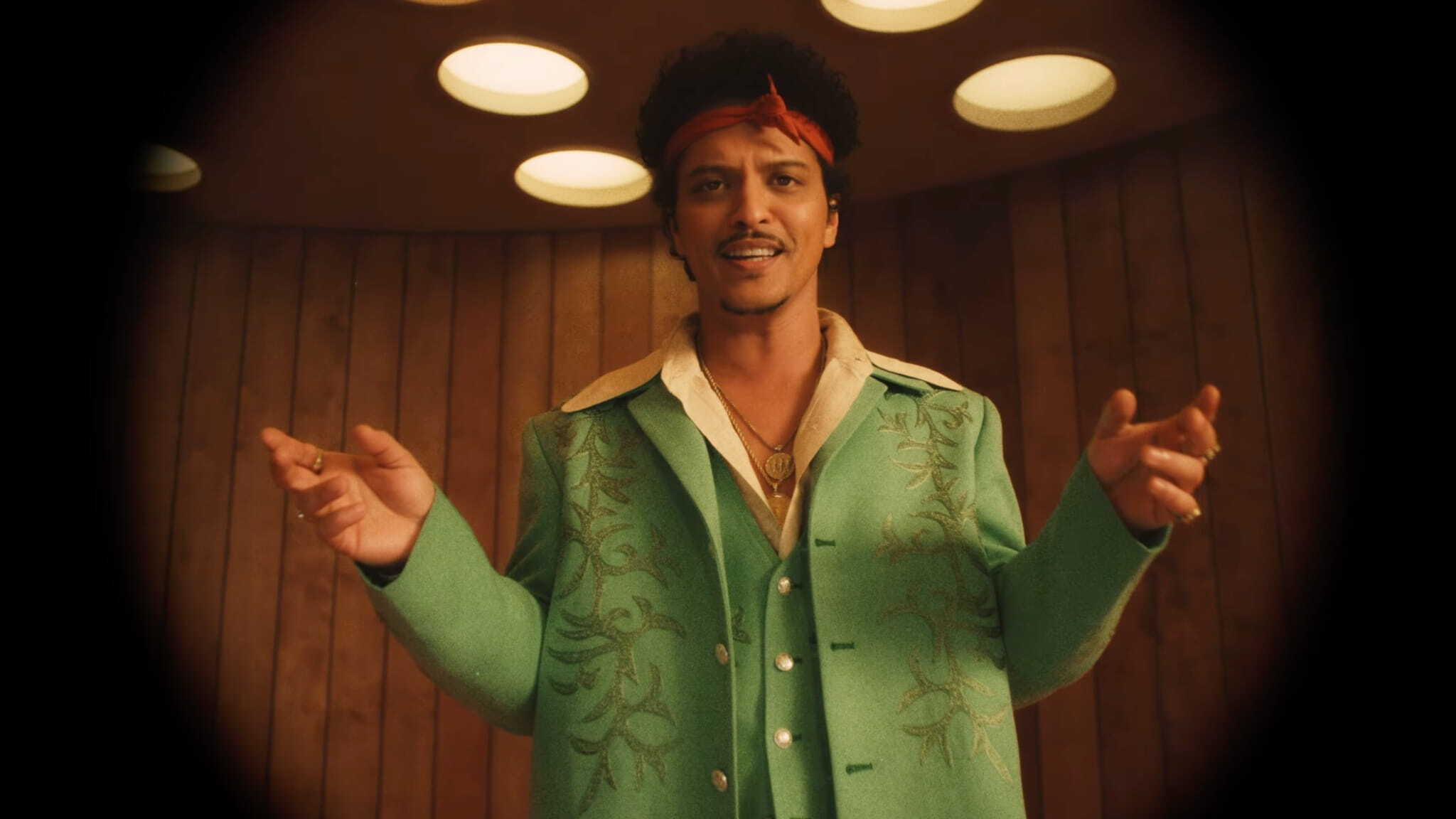 A man with curly hair and a red bandana, reminiscent of Bruno Mars, stands in a wood-paneled room with round ceiling lights, wearing a green embroidered jacket over a beige shirt and smiling as he holds his hands out.