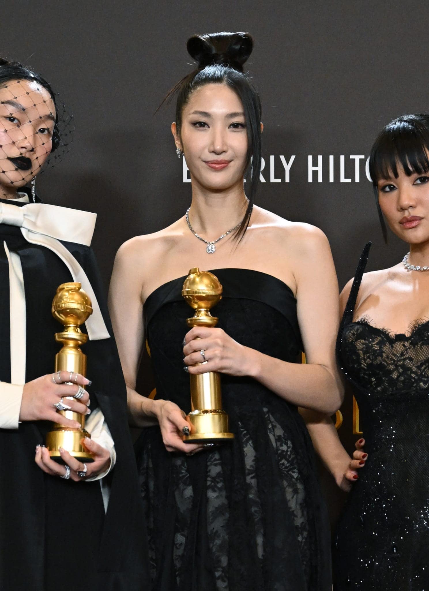 Three women in elegant black dresses stand together holding Golden Globe trophies, posing for a photo against a dark backdrop with event logos. One wears a dramatic veiled headpiece and large white bow.