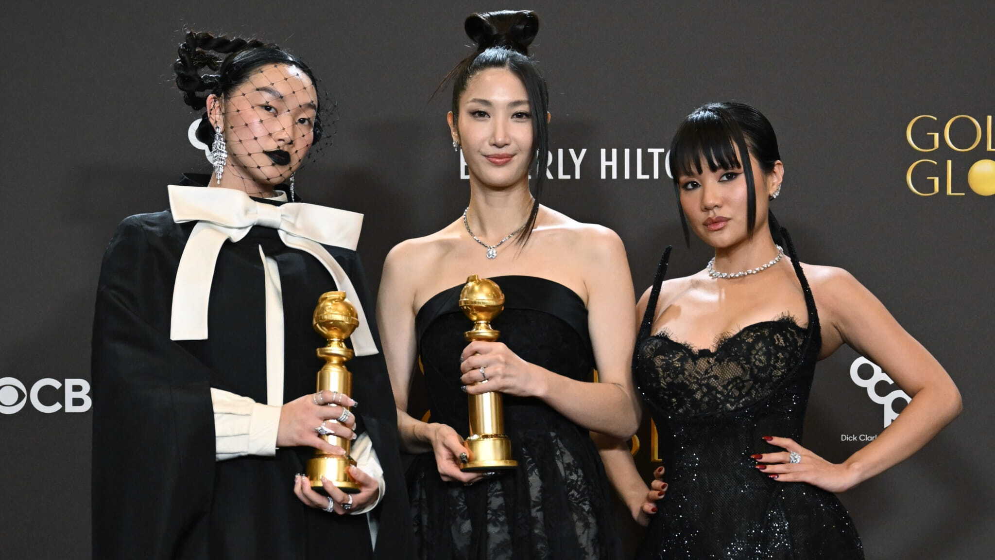 Three women in elegant black dresses stand together holding Golden Globe trophies, posing for a photo against a dark backdrop with event logos. One wears a dramatic veiled headpiece and large white bow.