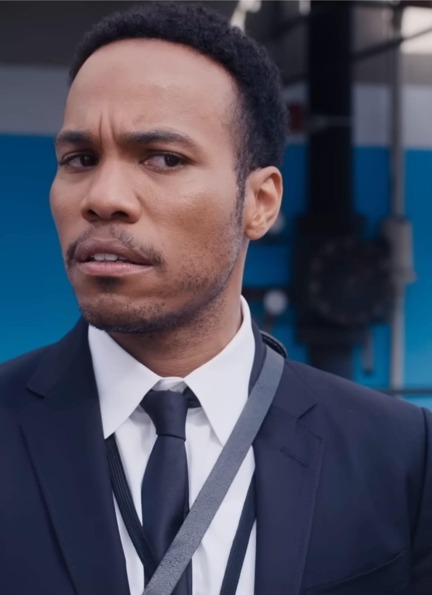A man in a suit with a striped tie and shoulder strap looks to the side with a skeptical or concerned expression. He stands outdoors in front of a blue and white background with metal railings.