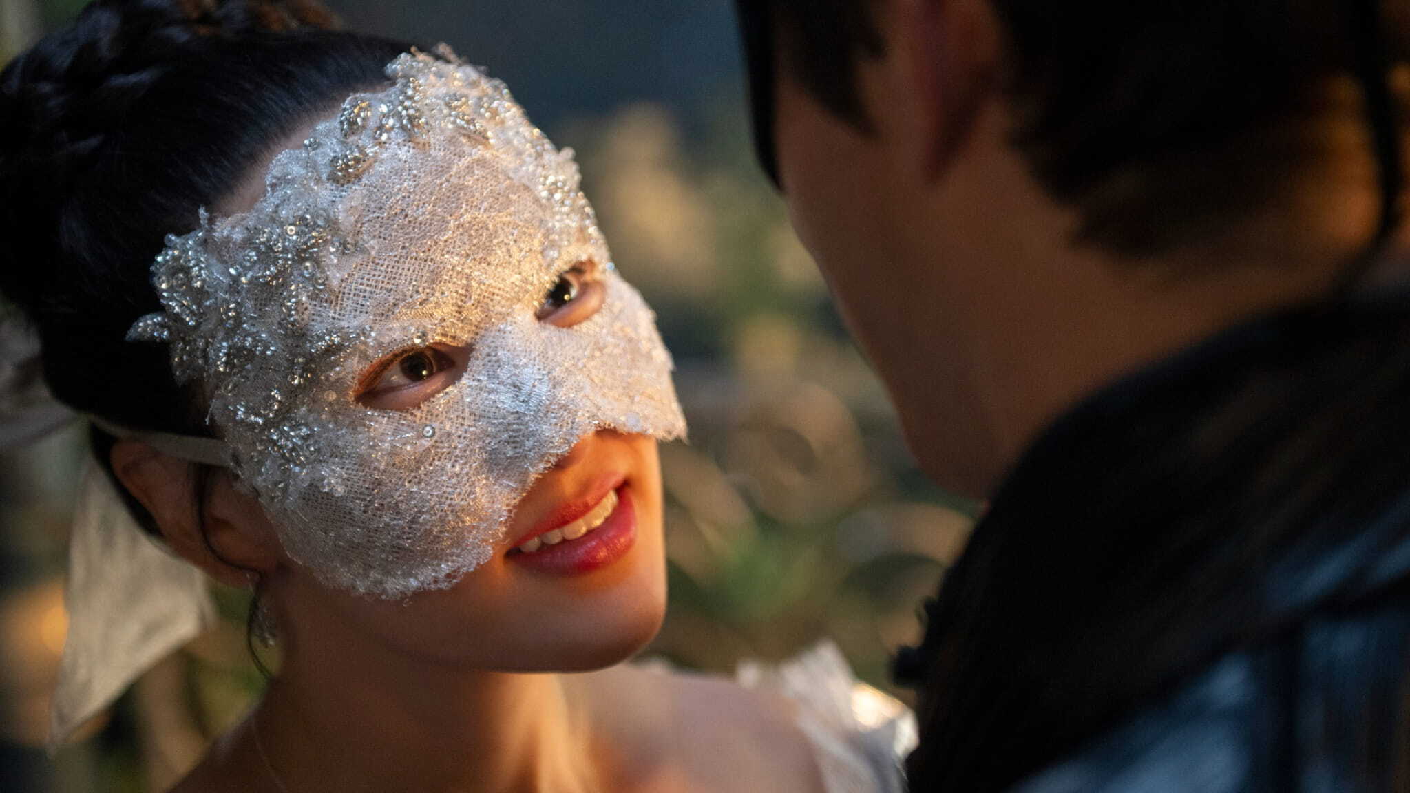 A woman wearing a white, lace, and beaded masquerade mask smiles warmly at a person facing her, who is also wearing a dark mask, in a softly lit setting.
