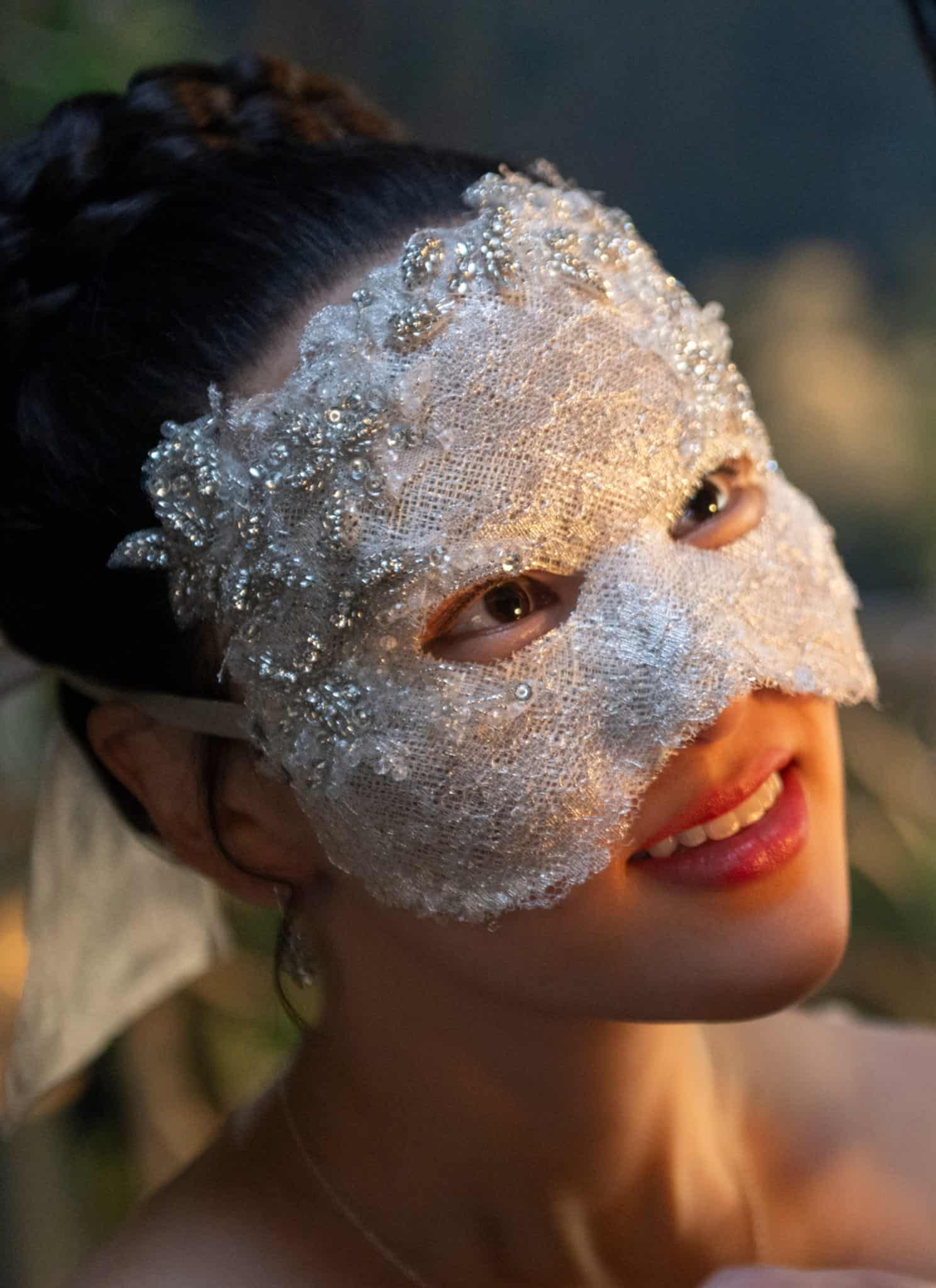 A woman wearing a white, lace, and beaded masquerade mask smiles warmly at a person facing her, who is also wearing a dark mask, in a softly lit setting.