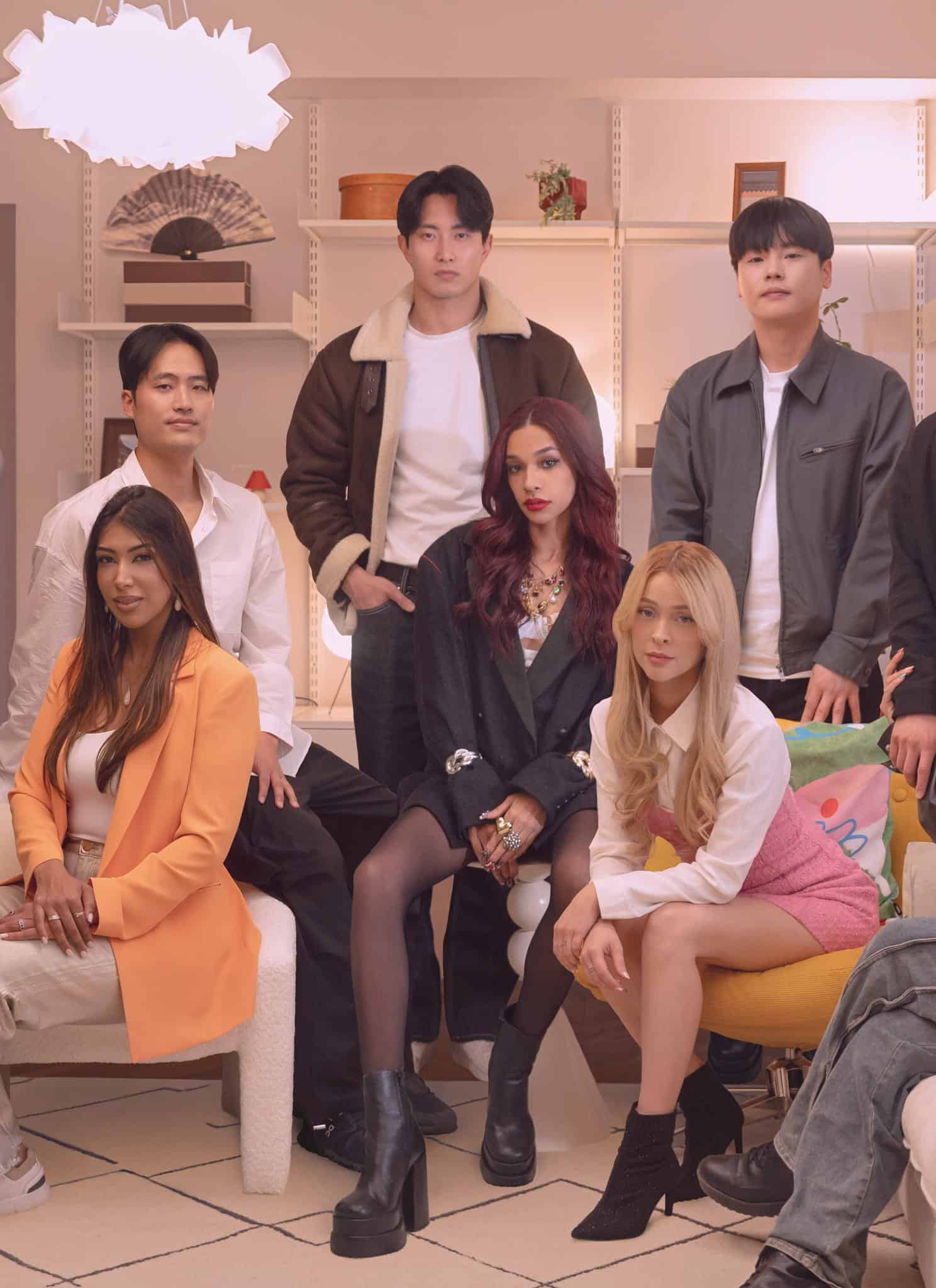 A group of eight stylish young adults, four men and four women, pose together in a modern, cozy living room with shelves, books, and warm lighting. Some are seated on chairs and a couch, while others stand behind them.