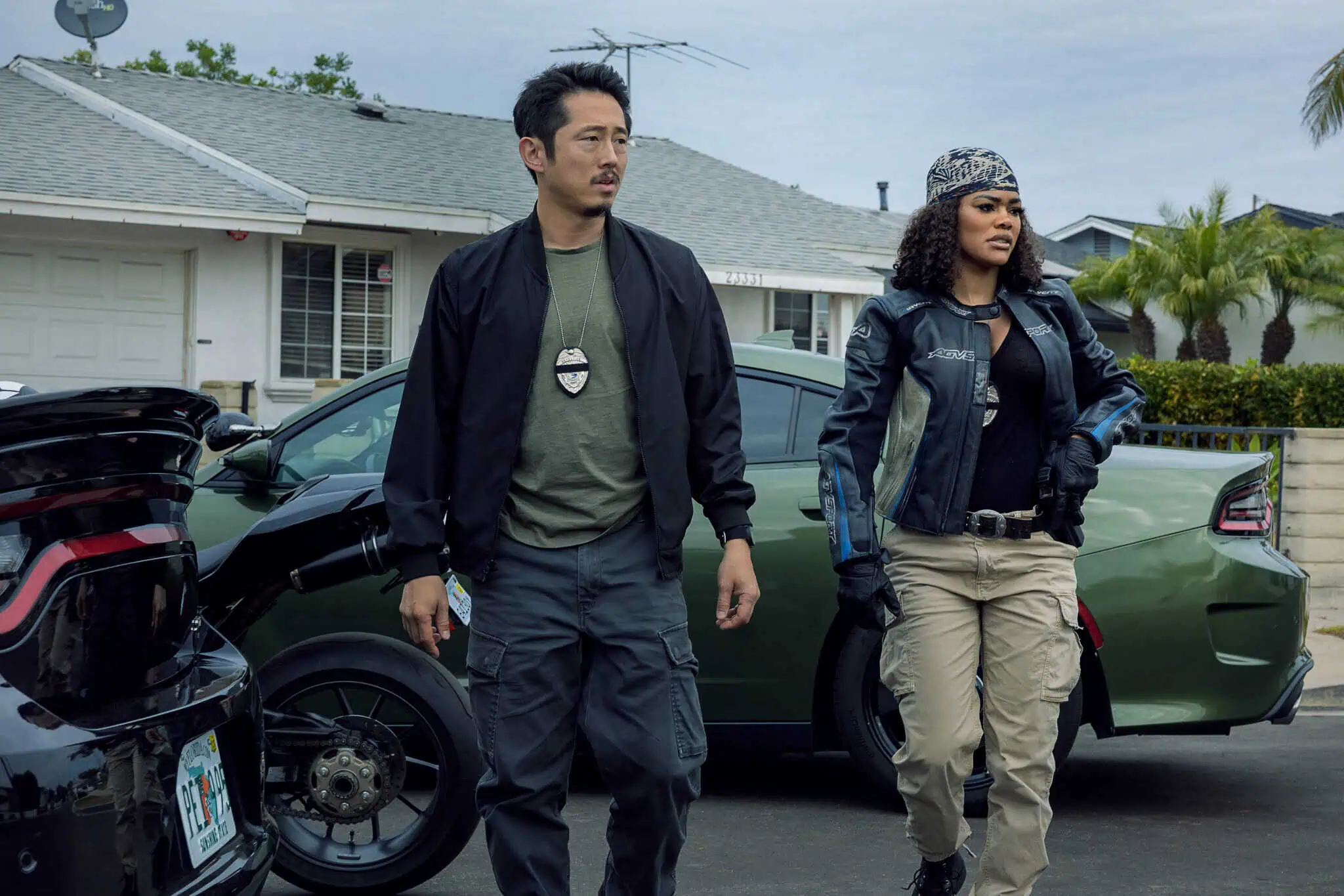 Two police officers stand beside parked cars in a suburban neighborhood, wearing tactical clothing and badges. One wears a black jacket and green shirt, the other has a beanie and blue jacket. Houses and palm trees are visible in the background.