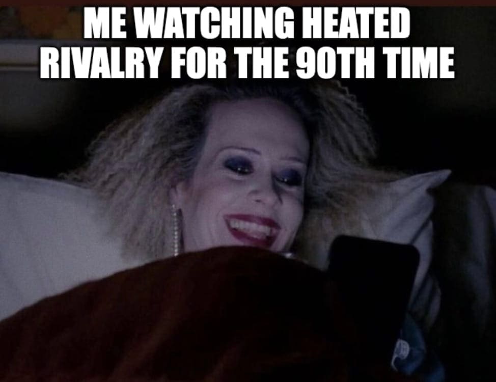 A person with frizzy blond hair and dramatic makeup smiles while lying in bed and looking at a phone. White text above reads: Me watching heated rivalry for the 90th time.
