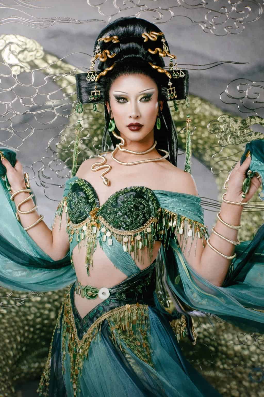 A person dressed in an elaborate, fantasy-inspired costume with green and gold details, jeweled accessories, and flowing sheer fabric, posing confidently against a shimmering, decorative background.