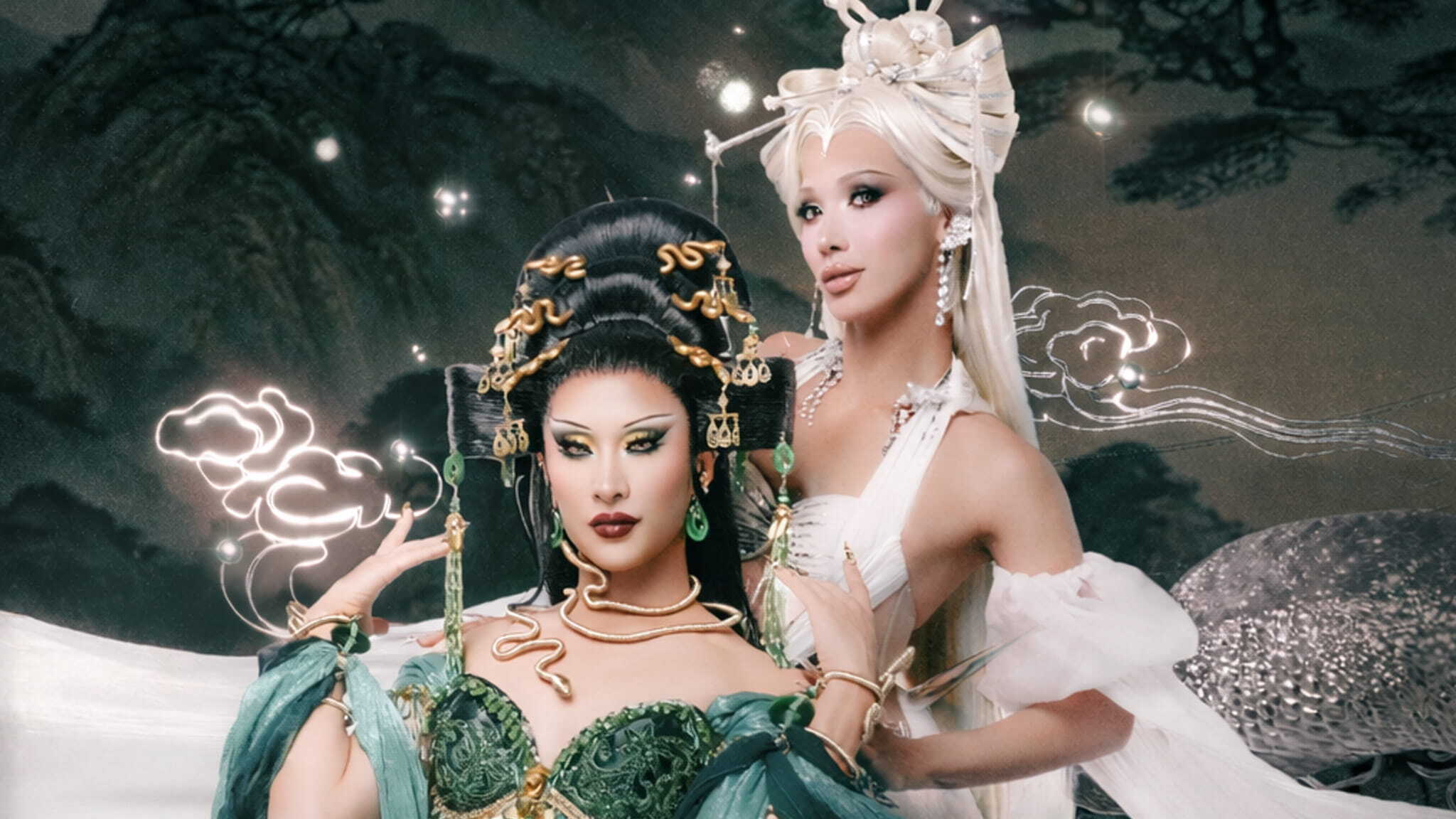 Two women in ornate fantasy costumes pose together; one wears a green dress with gold accessories and elaborate black hair, while the other wears white with long platinum hair and pearl details. The background features glowing clouds and nature.