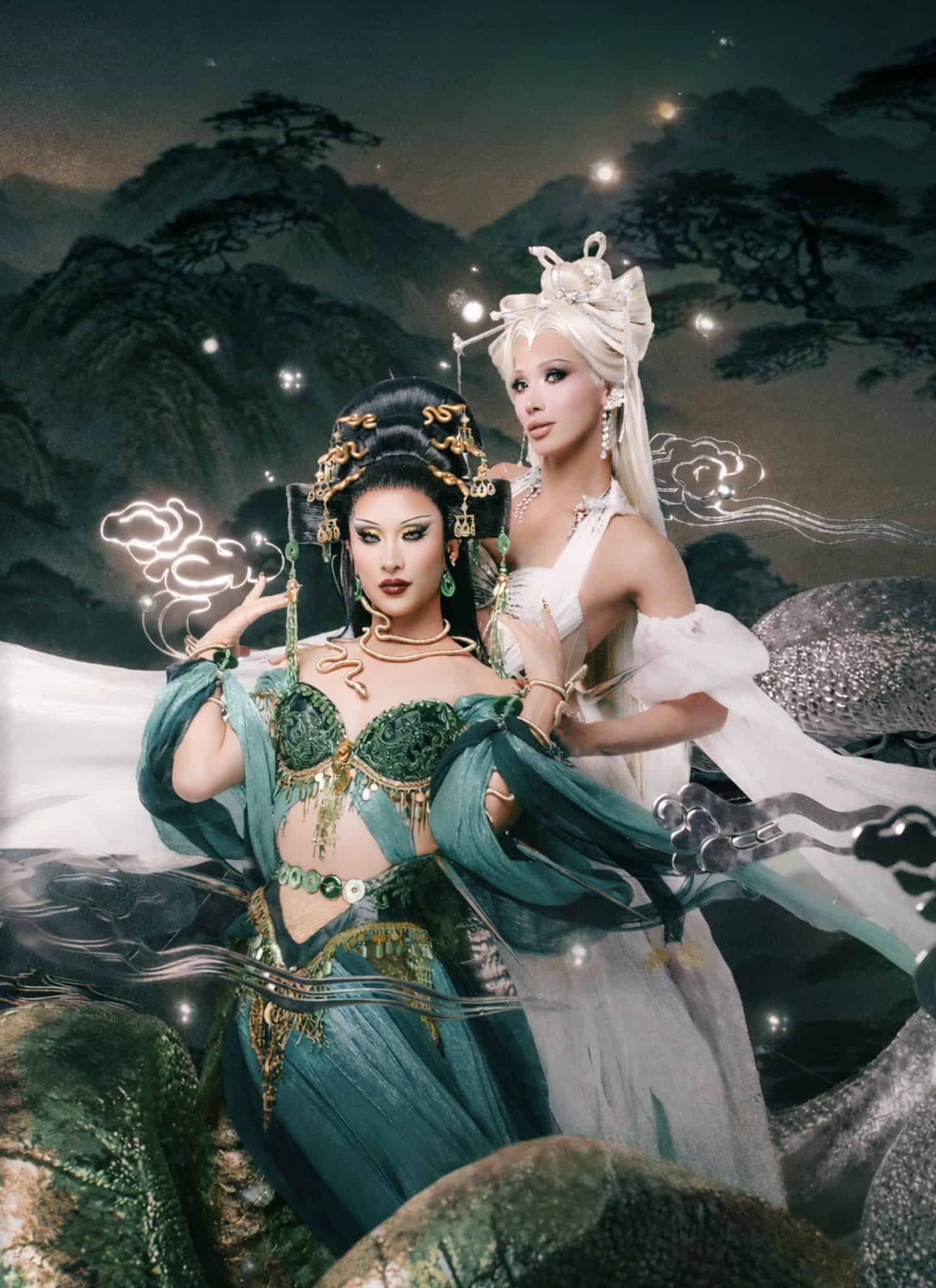 Two women in ornate fantasy costumes pose together; one wears a green dress with gold accessories and elaborate black hair, while the other wears white with long platinum hair and pearl details. The background features glowing clouds and nature.