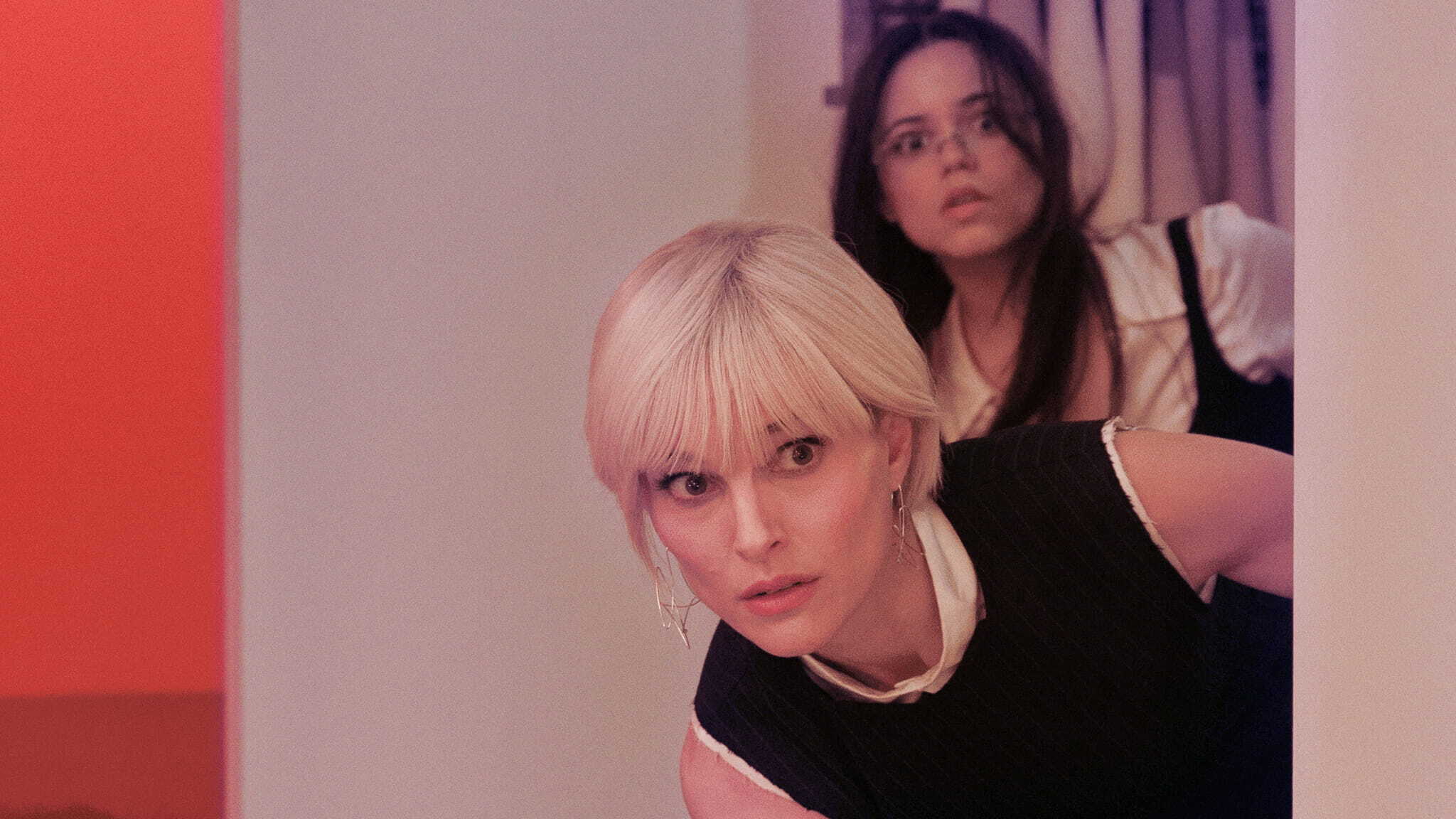 Two women peek around a corner with concerned expressions. The woman in front has short blonde hair and wears a sleeveless black top, while the woman behind has long dark hair, glasses, and a white shirt. The lighting is warm and soft.